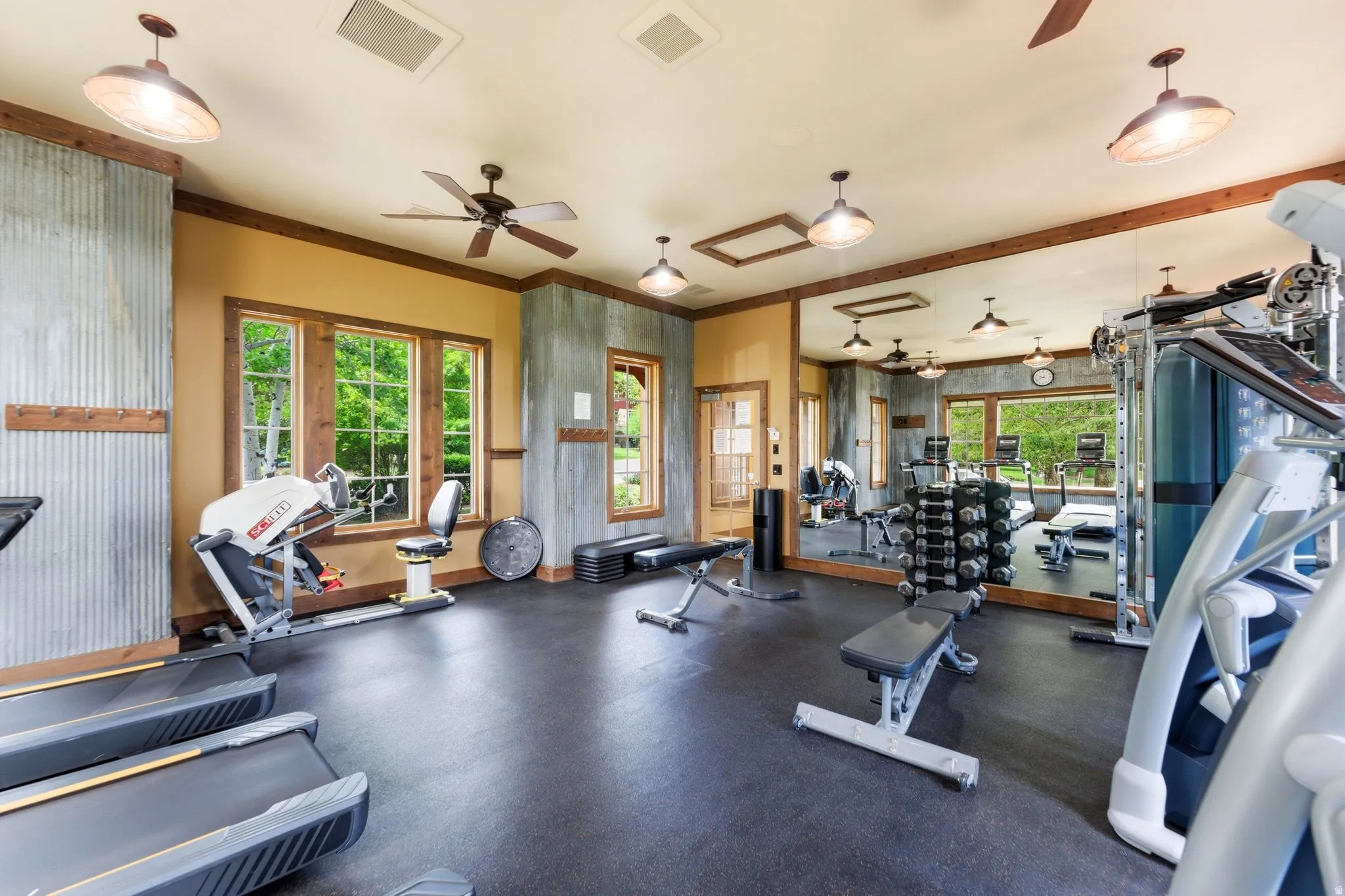 Dedicated fitness room featuring rubber flooring, multiple windows with wood-finish trim, a full-wall mirror, industrial-style pendant lighting, and ceiling fans