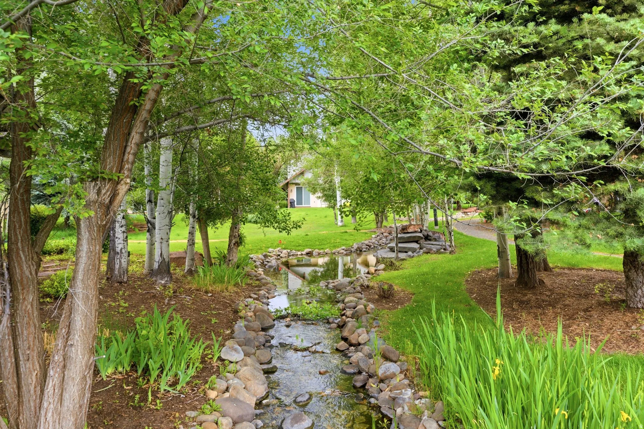 Landscaped grounds featuring a meandering stream with natural rock edging, verdant lawn, mature trees, and a flagstone pathway