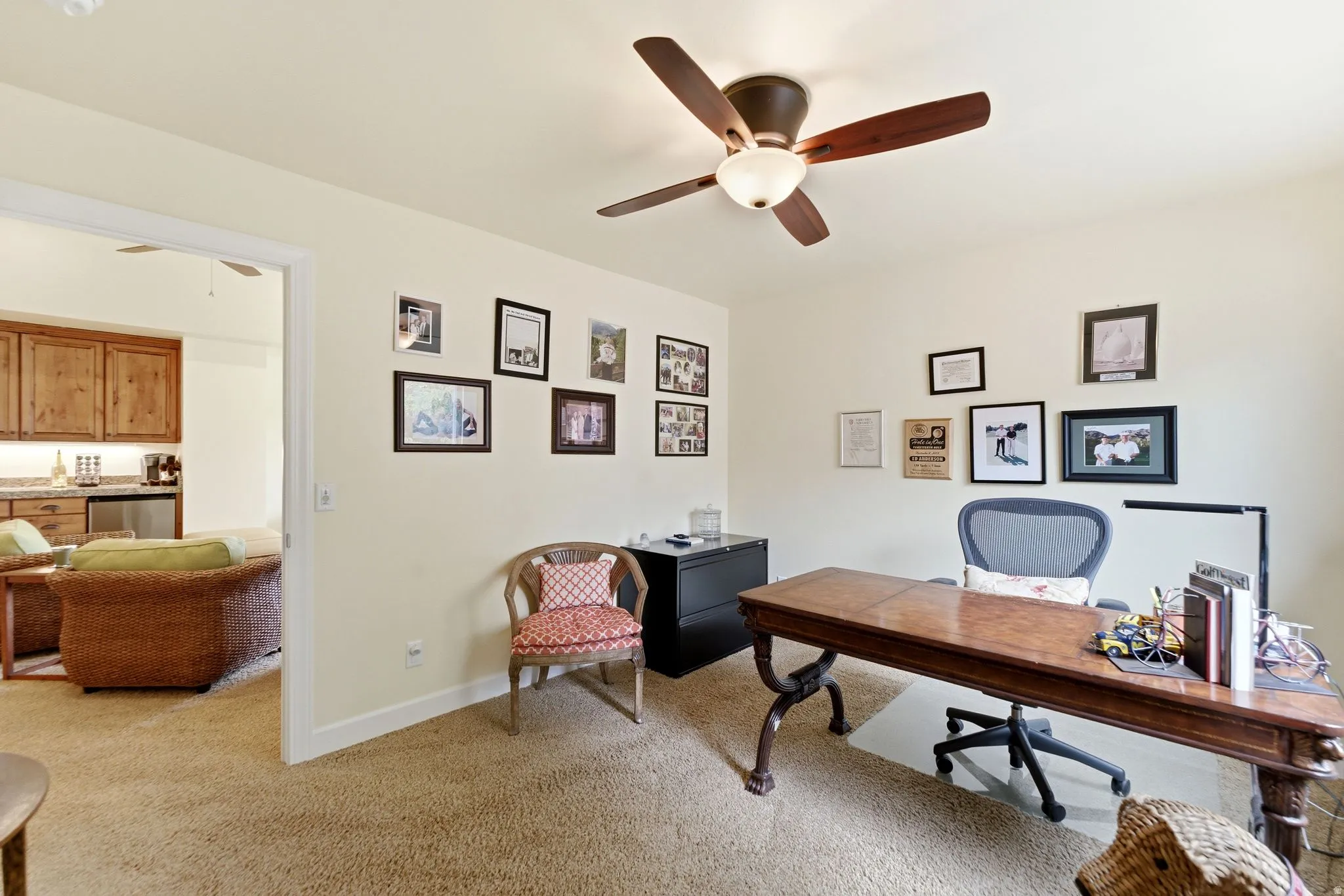 Warm-toned room featuring a ceiling fan with integrated lighting, textured carpet flooring, and an open doorway to an adjacent space with wood cabinetry and a countertop