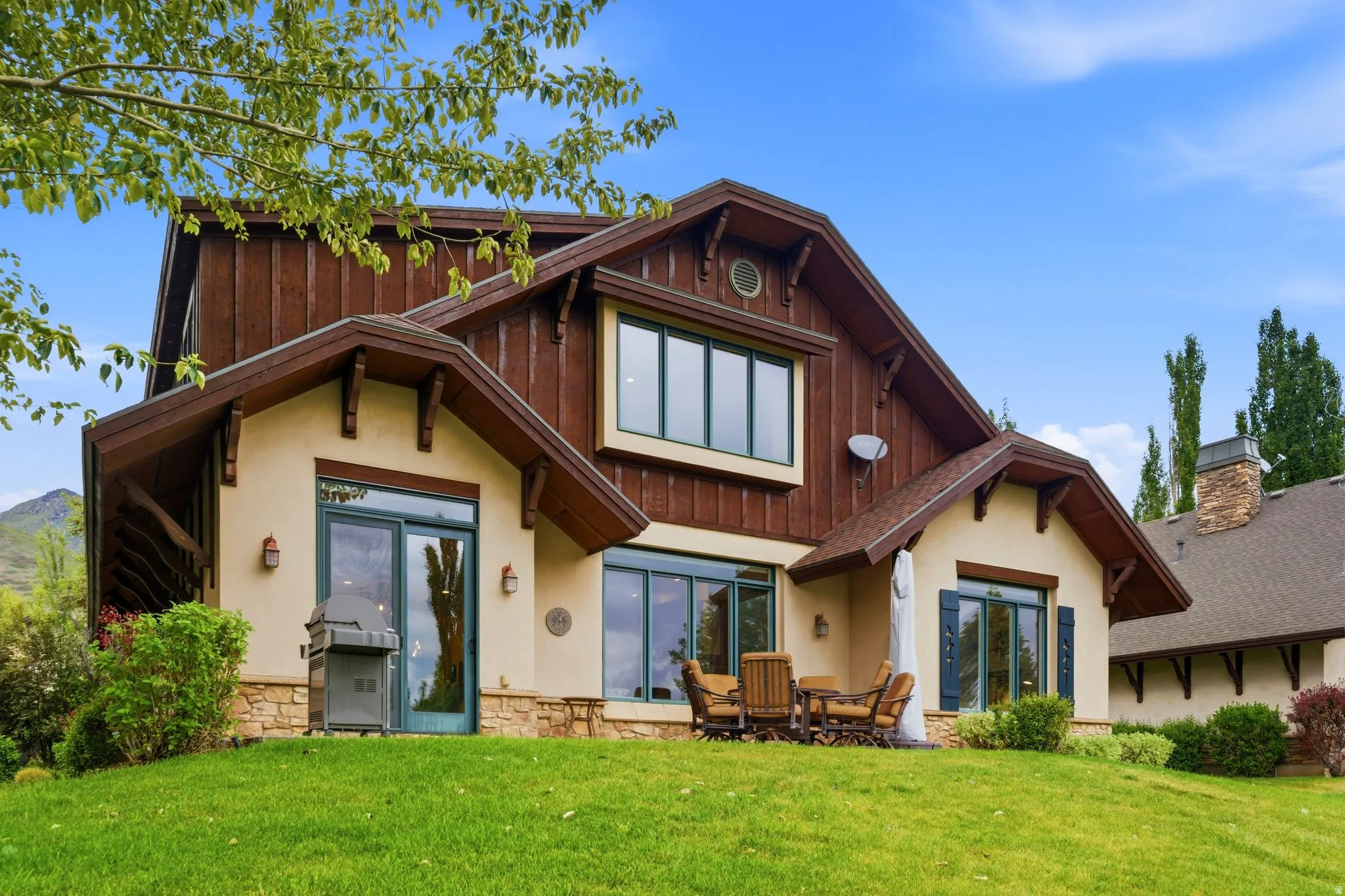 Exterior featuring a stucco and wood-paneled facade, stone accents, multiple large windows, a patio area, and a sloped lawn