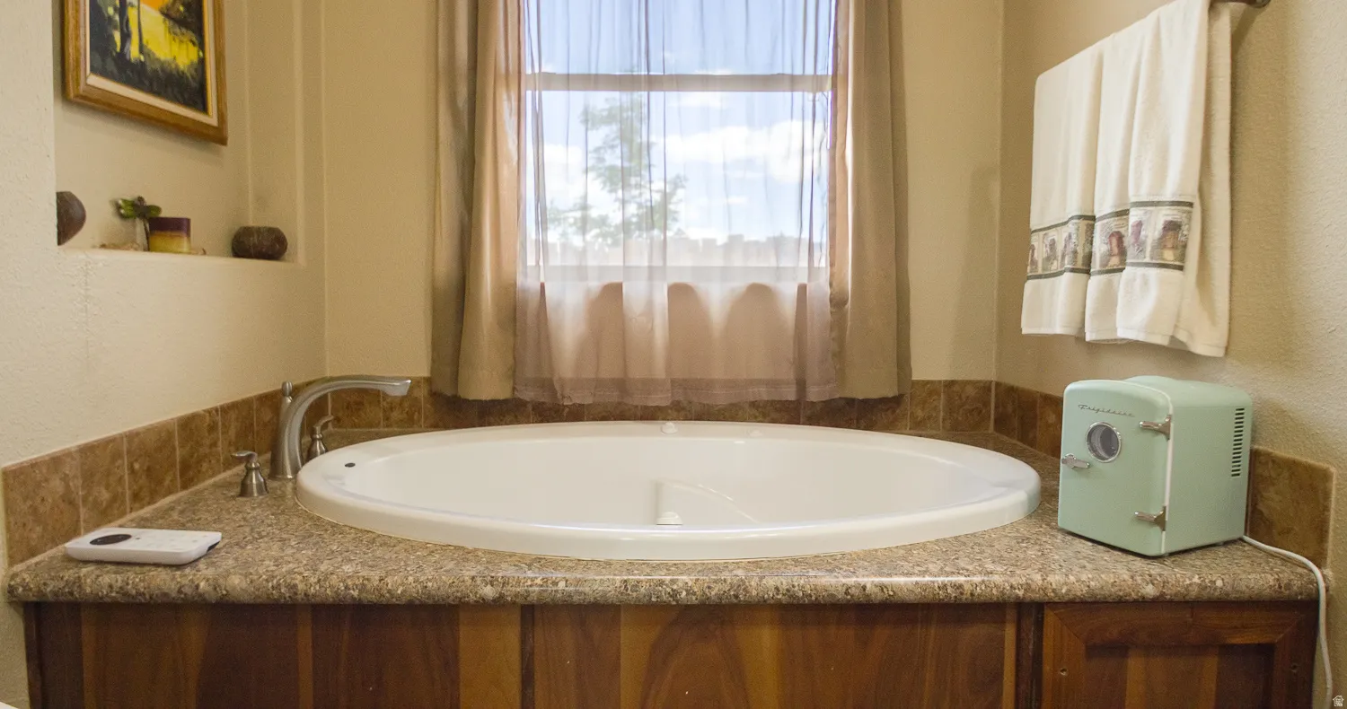 Built-in soaking tub with deck-mounted faucet, surrounded by wood-finish cabinetry, a stone-finish countertop, and stone-finish backsplash