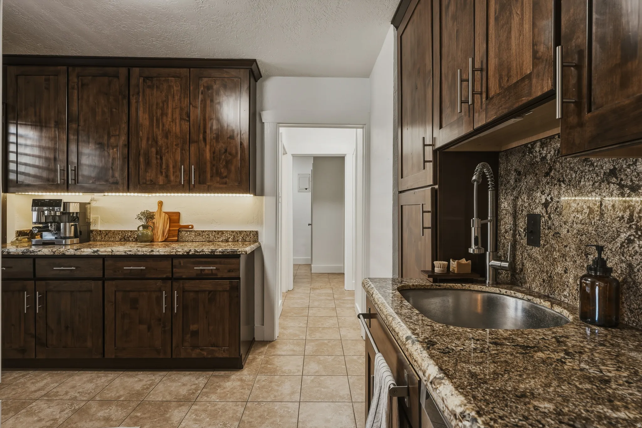 Kitchen featuring dark wood cabinetry, granite countertops, tile flooring, a stainless steel undermount sink, and under-cabinet lighting