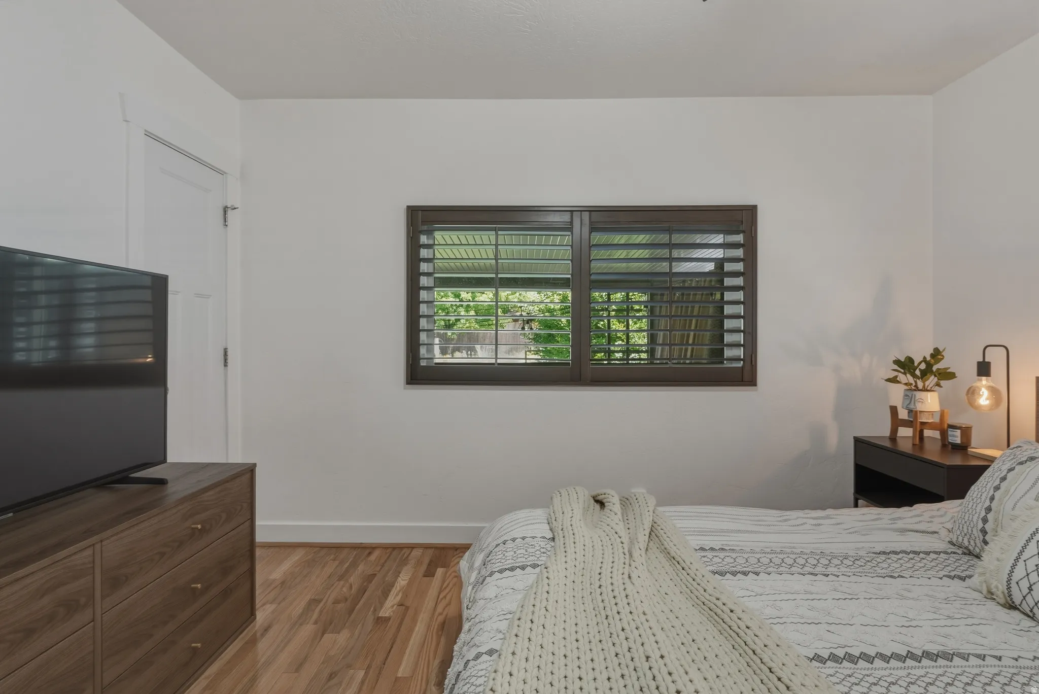 Room with hardwood flooring, white walls, and a window featuring dark wood plantation shutters