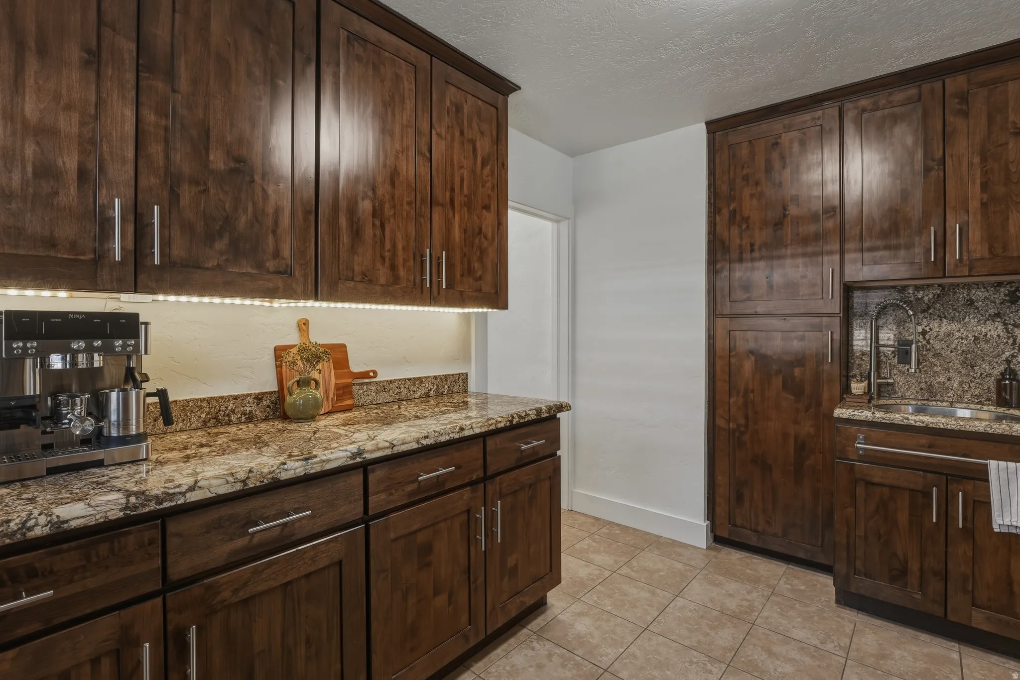 Kitchen featuring dark wood cabinetry, granite countertops, tile flooring, under-cabinet lighting, and a built-in sink with a gooseneck faucet