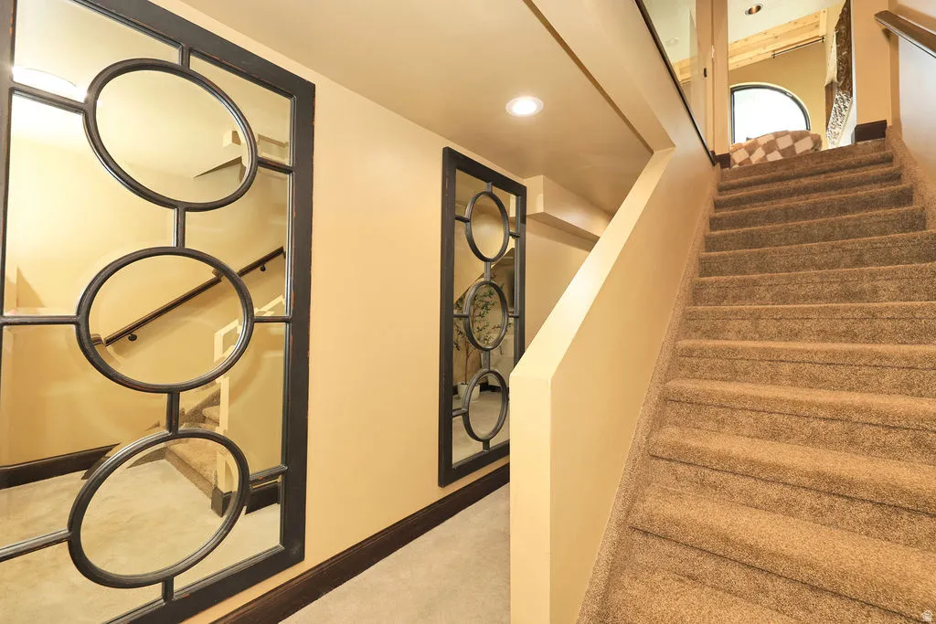 Carpeted staircase leading to walk out basement with recessed lighting and a decorative wall mirror featuring circular elements