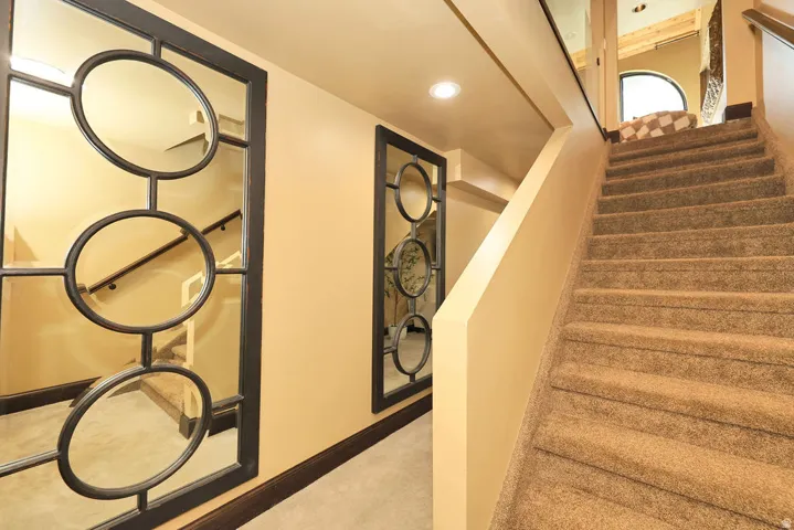 Carpeted staircase leading to walk out basement with recessed lighting and a decorative wall mirror featuring circular elements