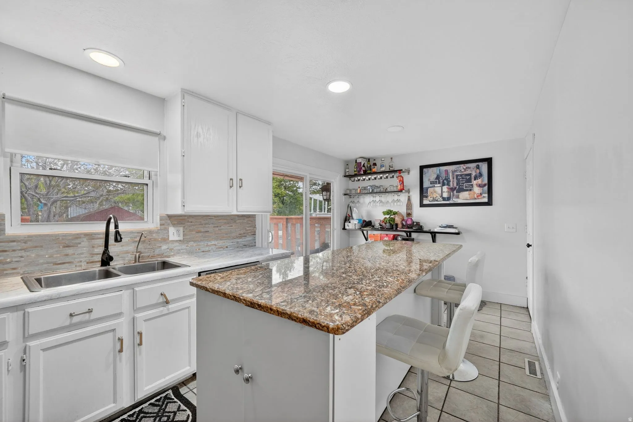 Kitchen featuring white cabinetry, a dual basin stainless steel sink with a matte black faucet, a textured tile backsplash, and a breakfast bar with a stone countertop