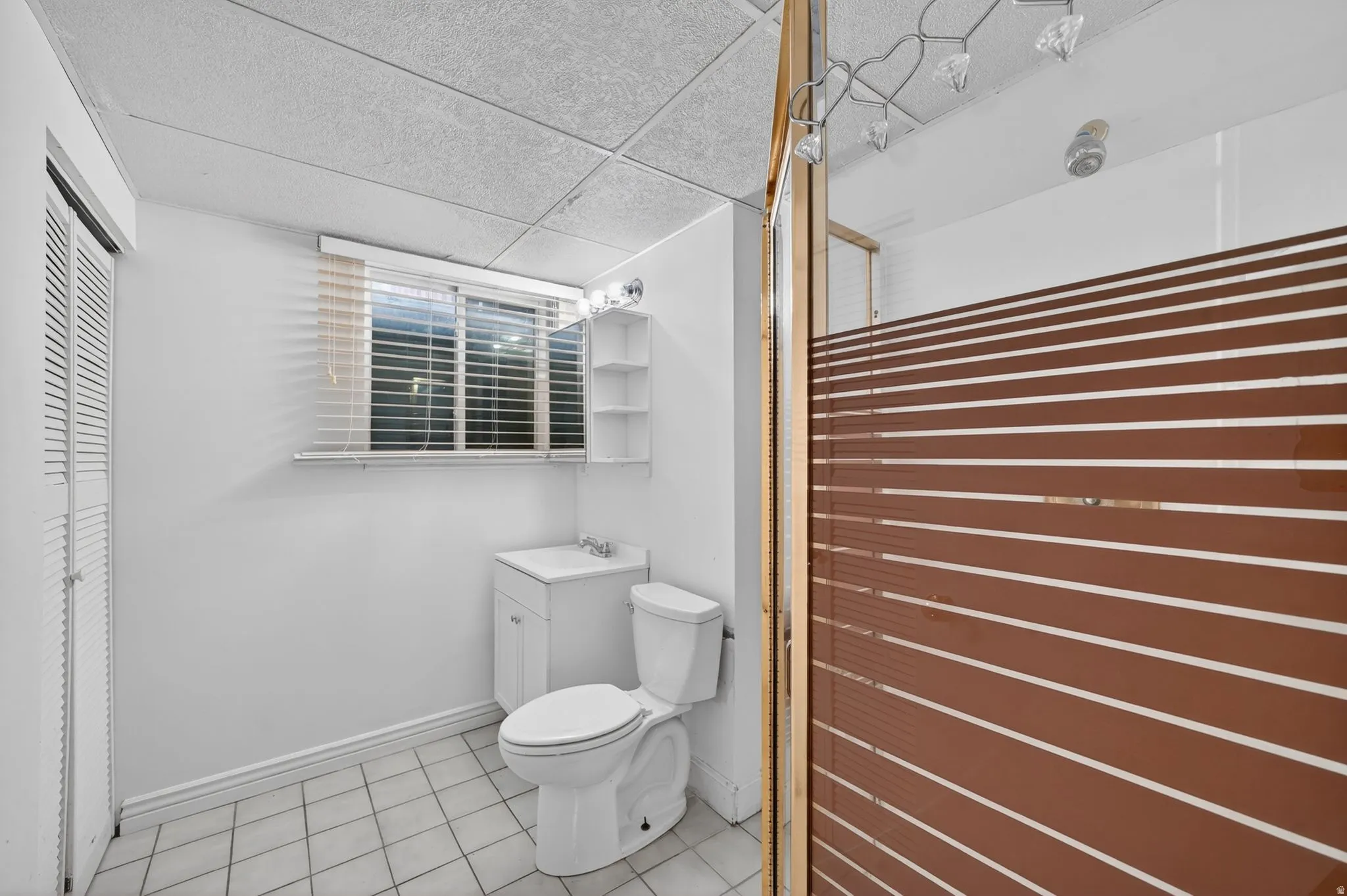 Bathroom featuring a single window with blinds, white vanity with integrated sink, toilet, tiled flooring, and a drop ceiling