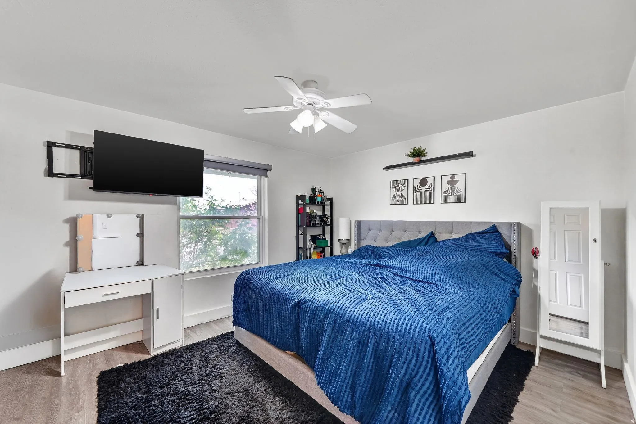 Bedroom featuring wood-finish flooring, a ceiling fan, a window with roller shades, and a wall-mounted television