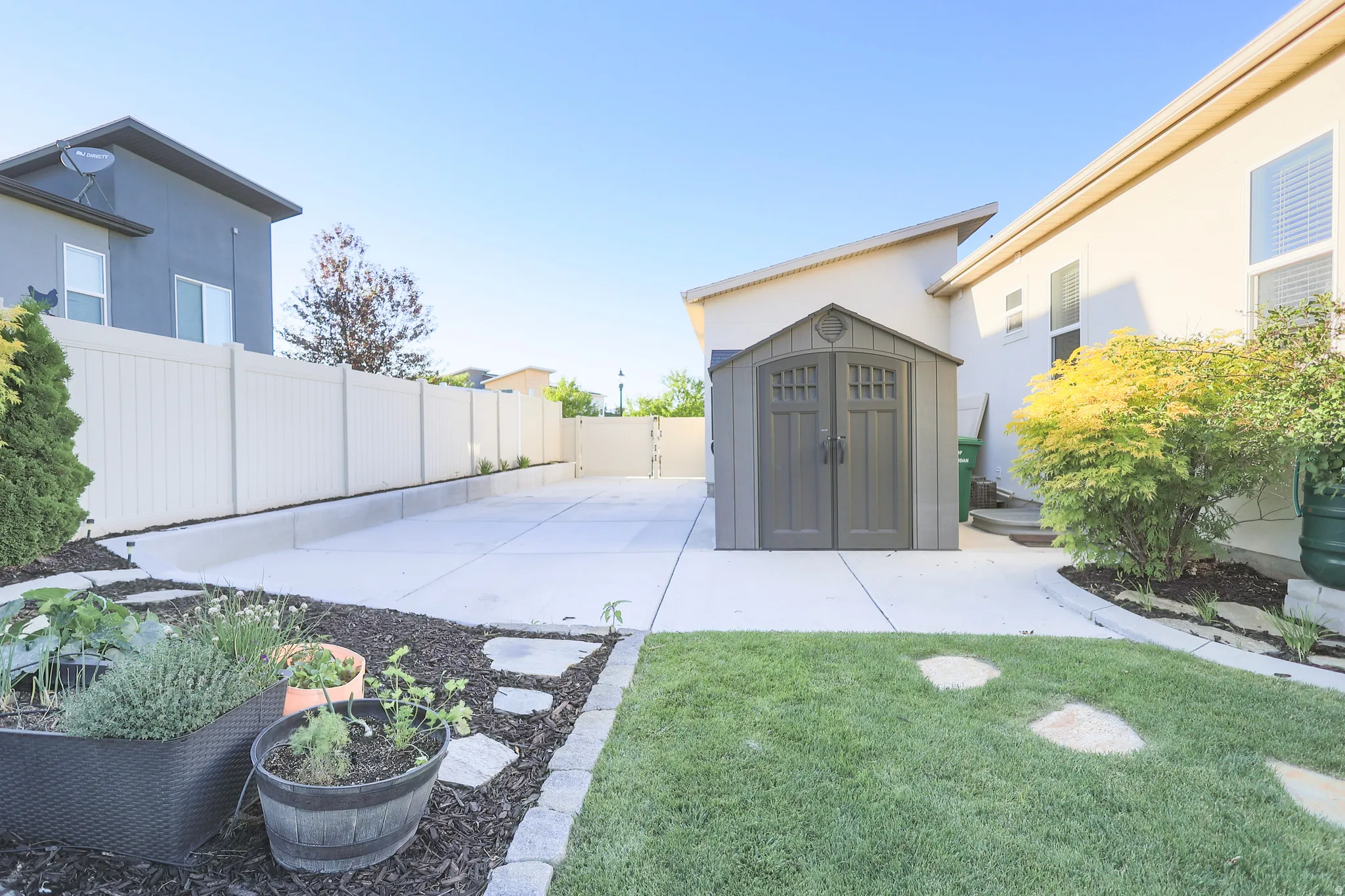 Expansive concrete patio area featuring a detached storage shed, a manicured lawn section, and established perimeter plantings/ gardening