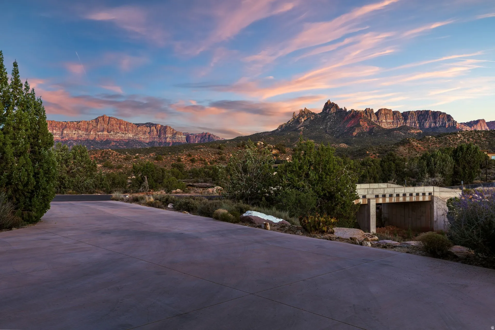 Expansive paved driveway bordered by mature evergreen trees and arid landscaping