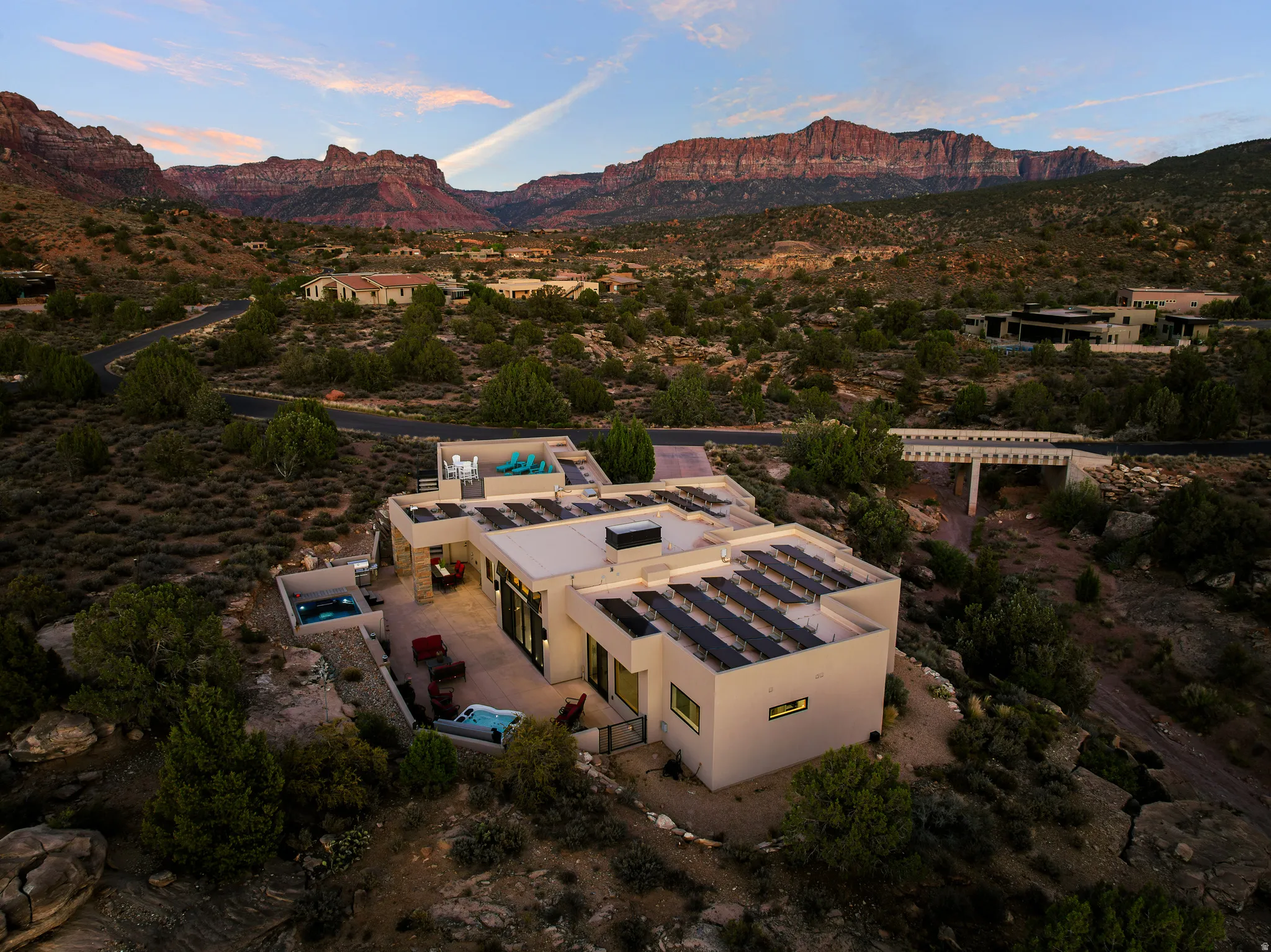 Elevated dusk view capturing the home's footprint, rooftop solar array, and rear outdoor living with the swim spa and cliff backdrop beyond.