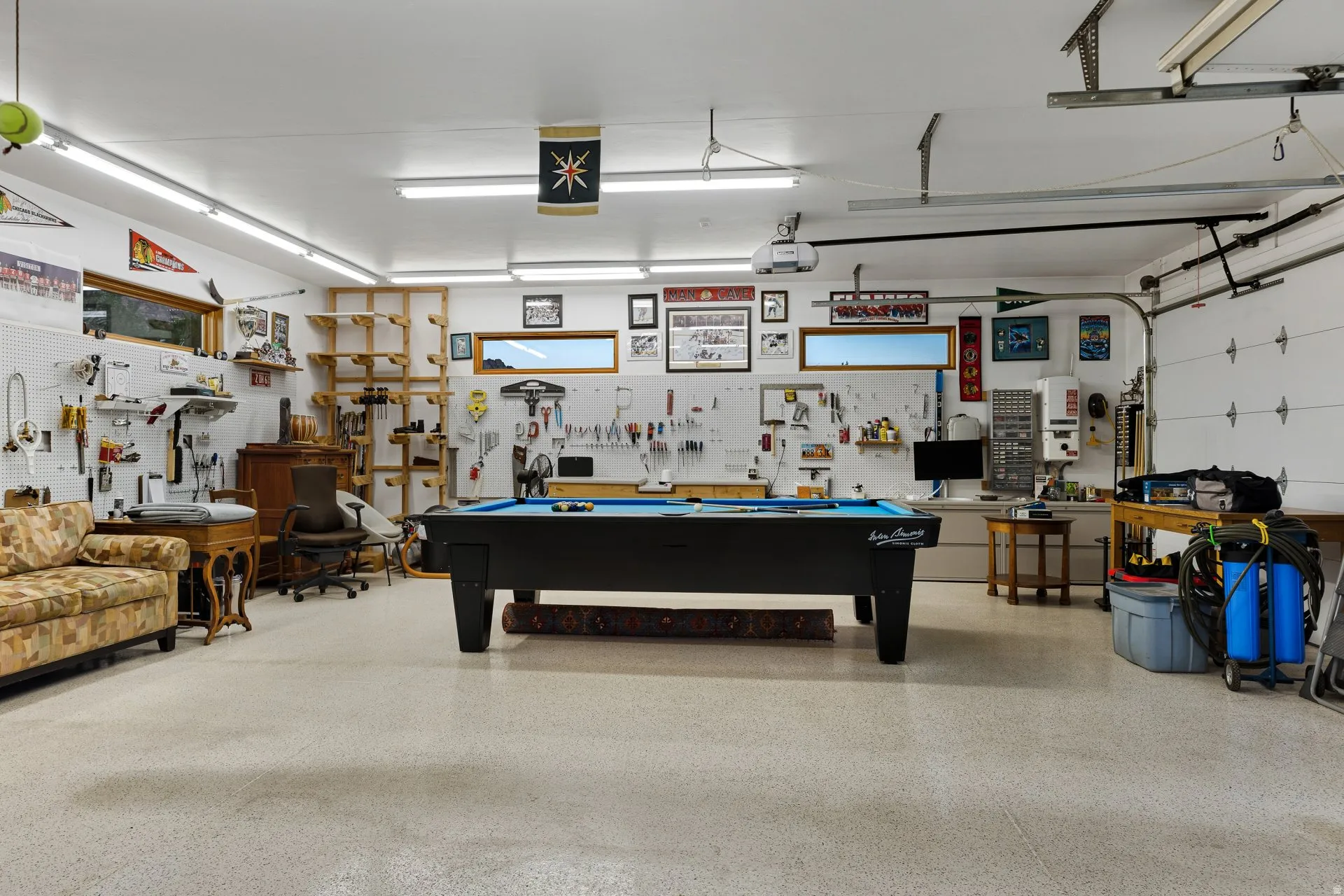 Spacious garage with light-colored epoxy flooring, featuring a dedicated workspace with built-in shelving, multiple horizontal windows providing natural light, and overhead fluorescent lighting