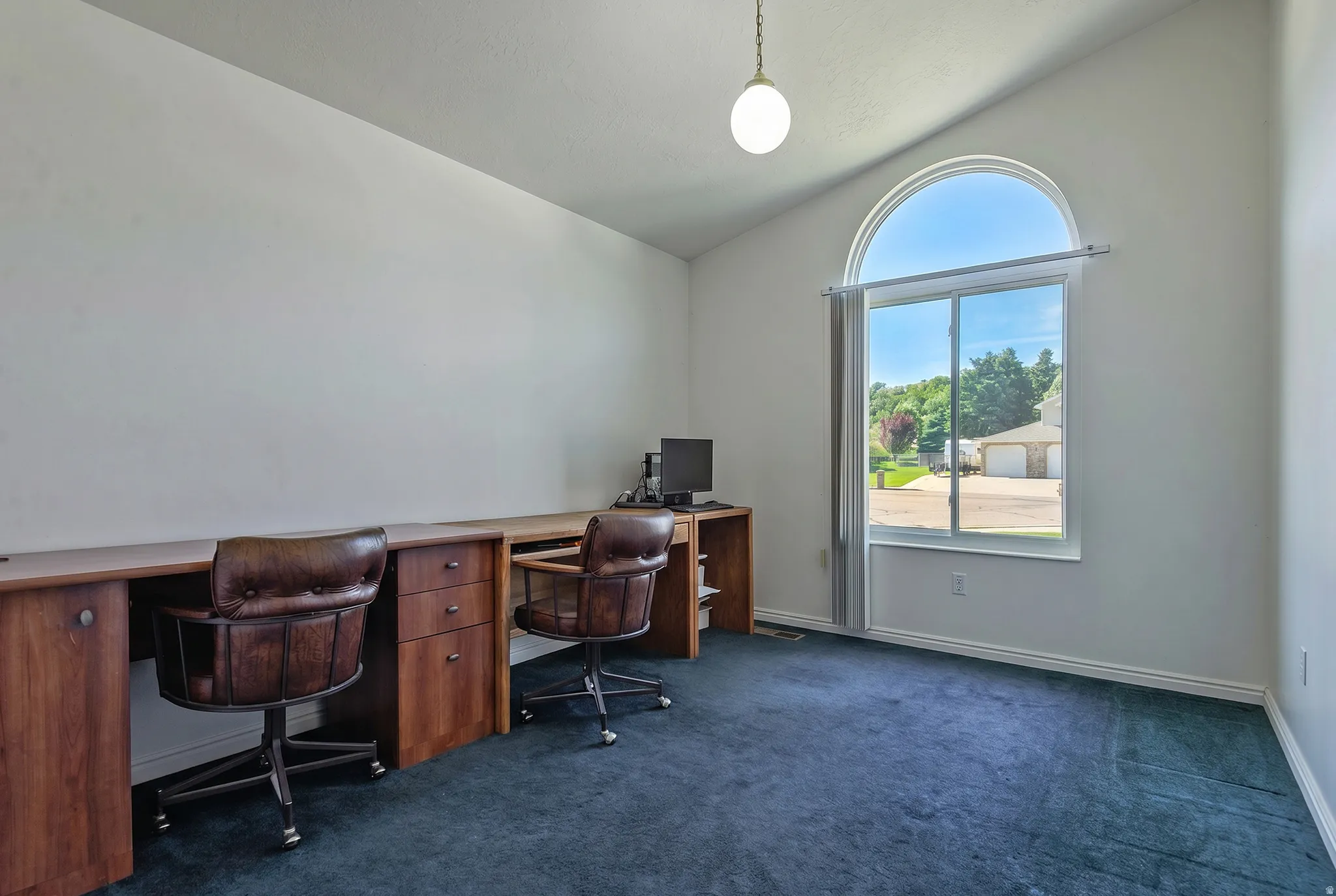 Vaulted ceiling room featuring a large arched window, built-in wooden desk with storage, and blue carpeting