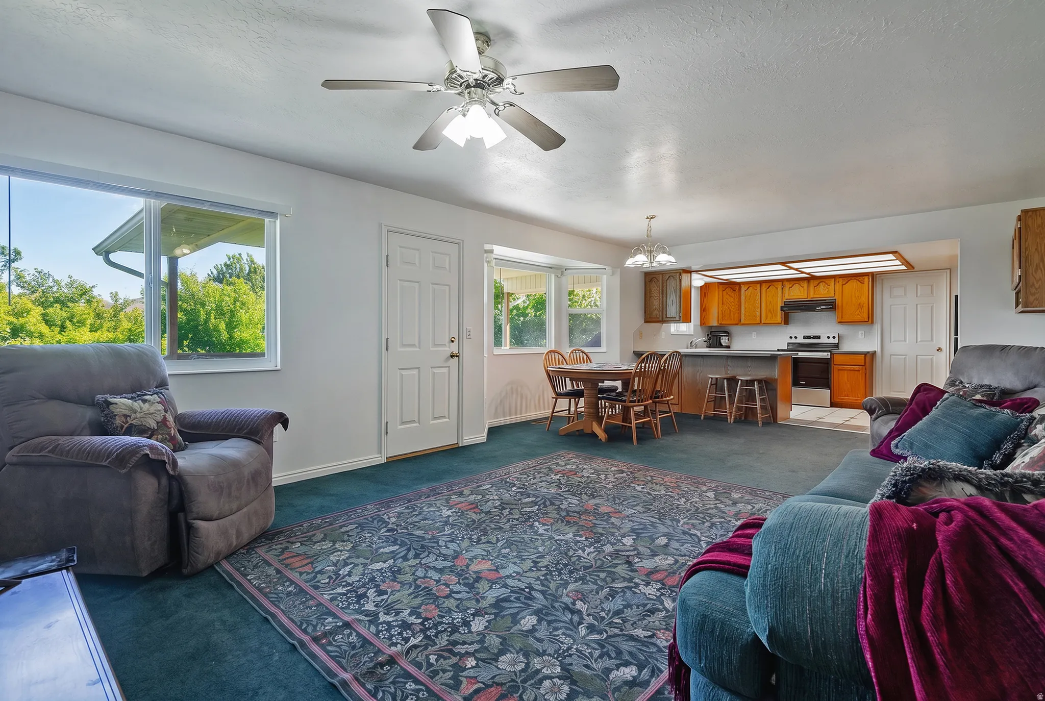 Open-concept living area featuring a ceiling fan, broad windows, and a carpeted floor