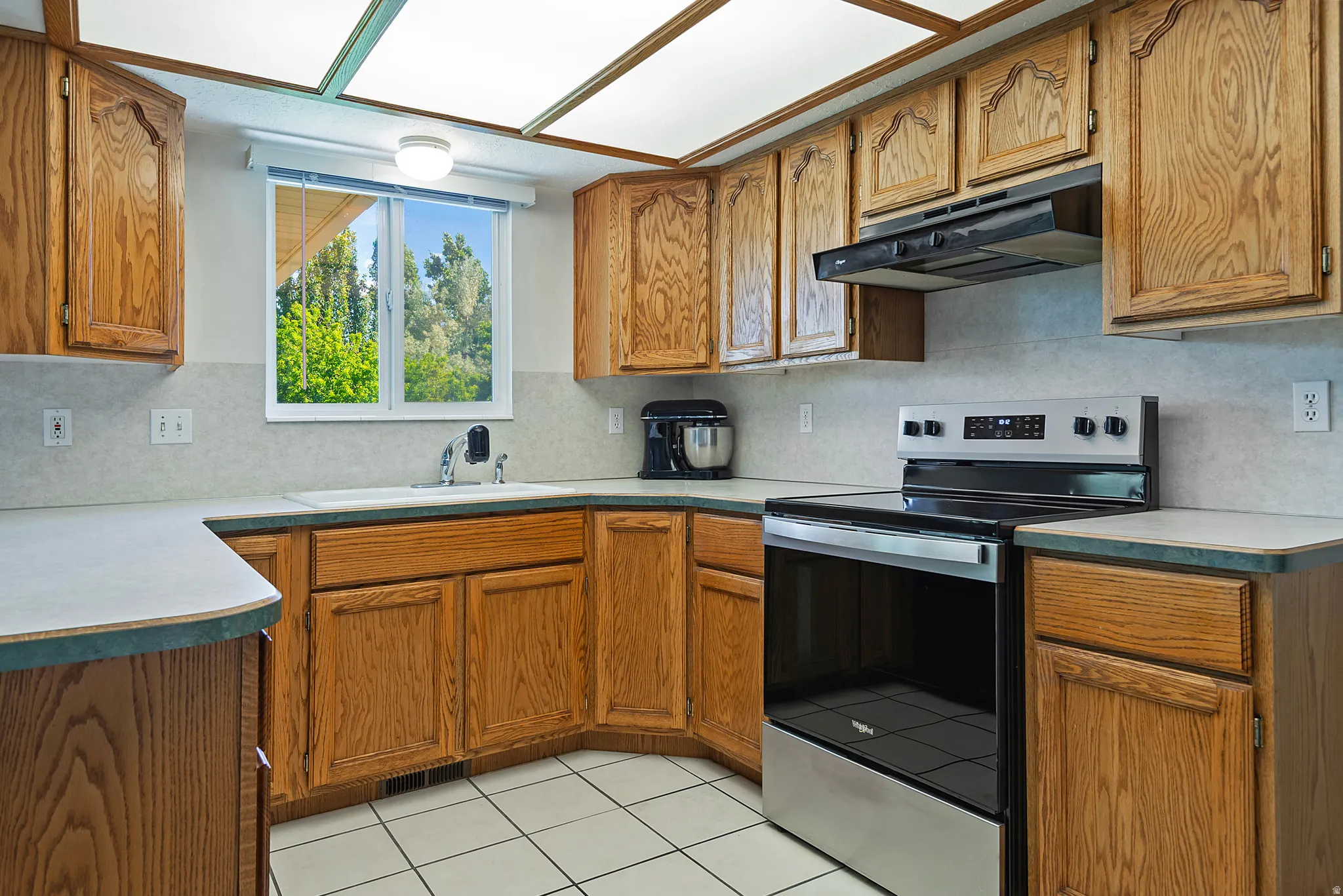 Kitchen featuring wood cabinetry, white tile flooring, stainless steel electric range, and a window overlooking exterior greenery