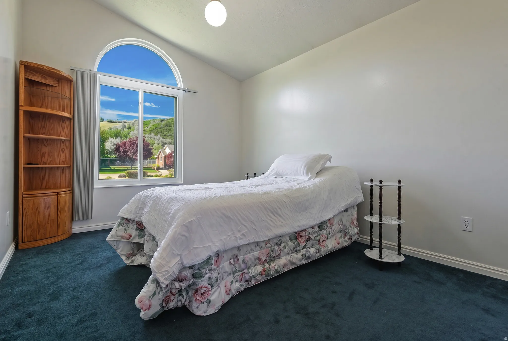Vaulted ceiling room featuring a large arched window, a corner wood-finish bookshelf with cabinet storage, and dark green carpeting