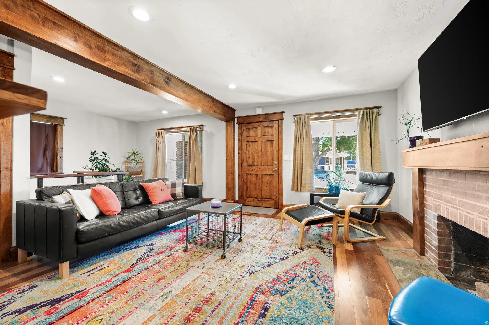 Living space featuring large southern windows, exposed wood beam, recessed lighting, wood flooring, a wood-panel entry door, and a brick fireplace with a wood mantle