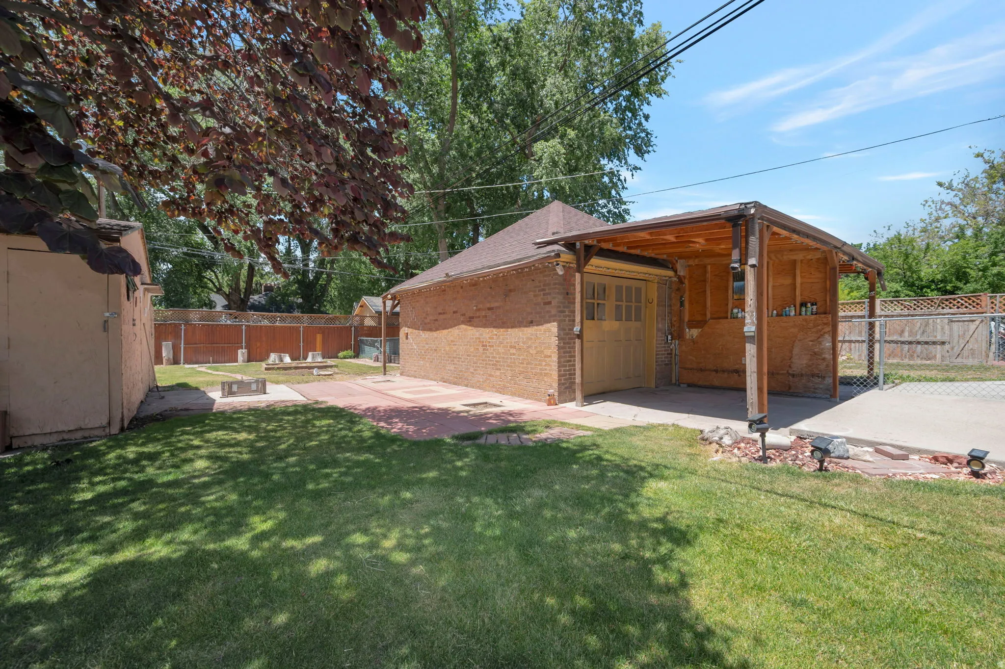 Expansive backyard featuring a brick garage structure, a wood-covered patio area, a concrete slab, a brick-paved patio, and a separate shed building