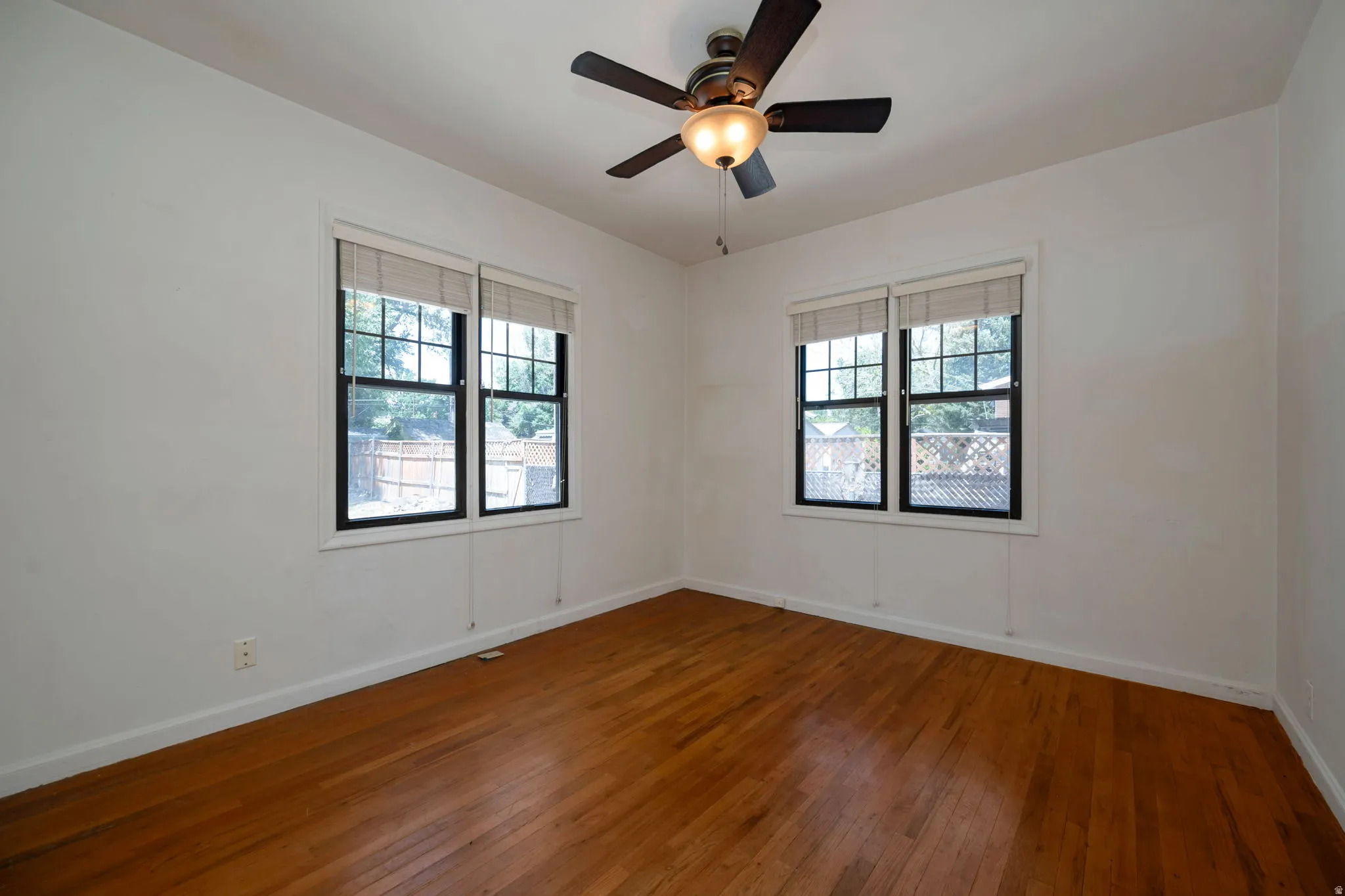 Hardwood flooring extends throughout the room, complemented by two windows with blinds and a ceiling fan with integrated lighting