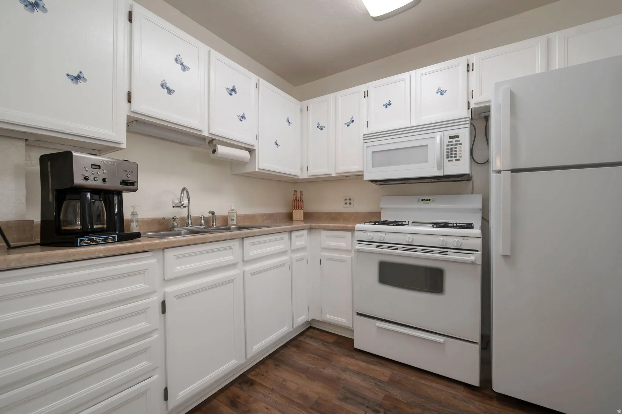 Kitchen featuring white cabinetry, light-toned countertops, a stainless steel sink, wood-finish flooring, and white appliances including a refrigerator, range, and microwave