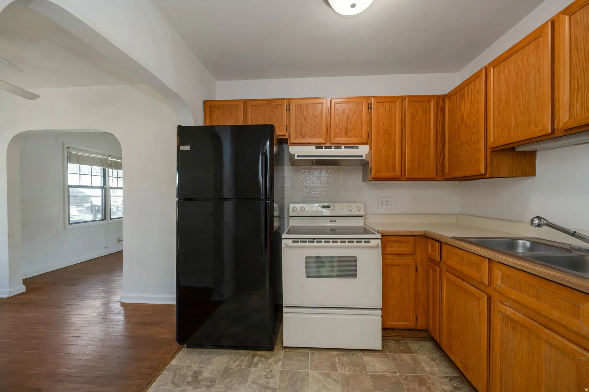 Kitchen featuring wood cabinetry, white appliances, a double basin sink, and an arched doorway opening to a room with wood-finish flooring and windows