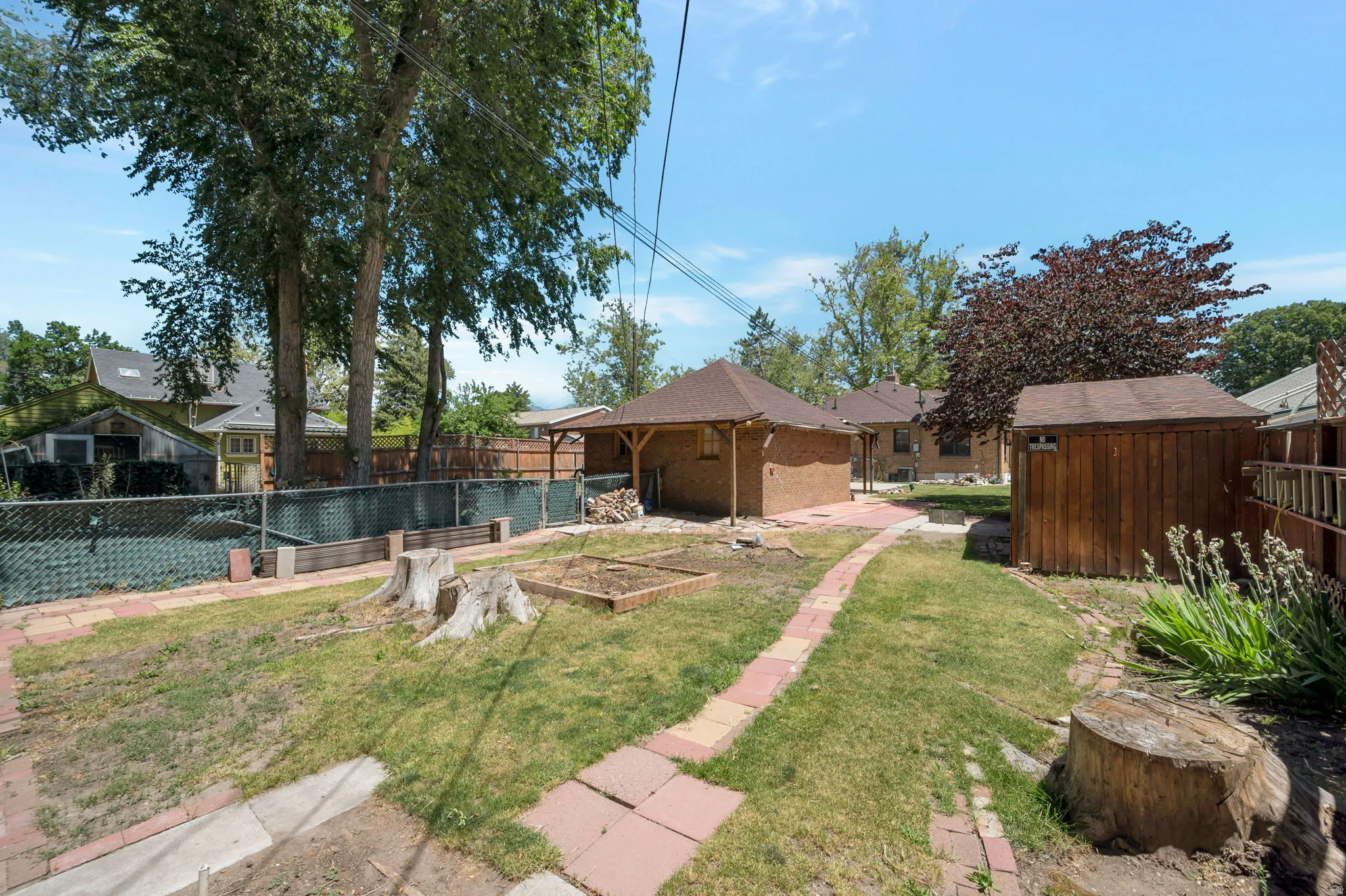Spacious backyard featuring a brick paver pathway, mature trees, a wooden storage shed, and a covered patio structure