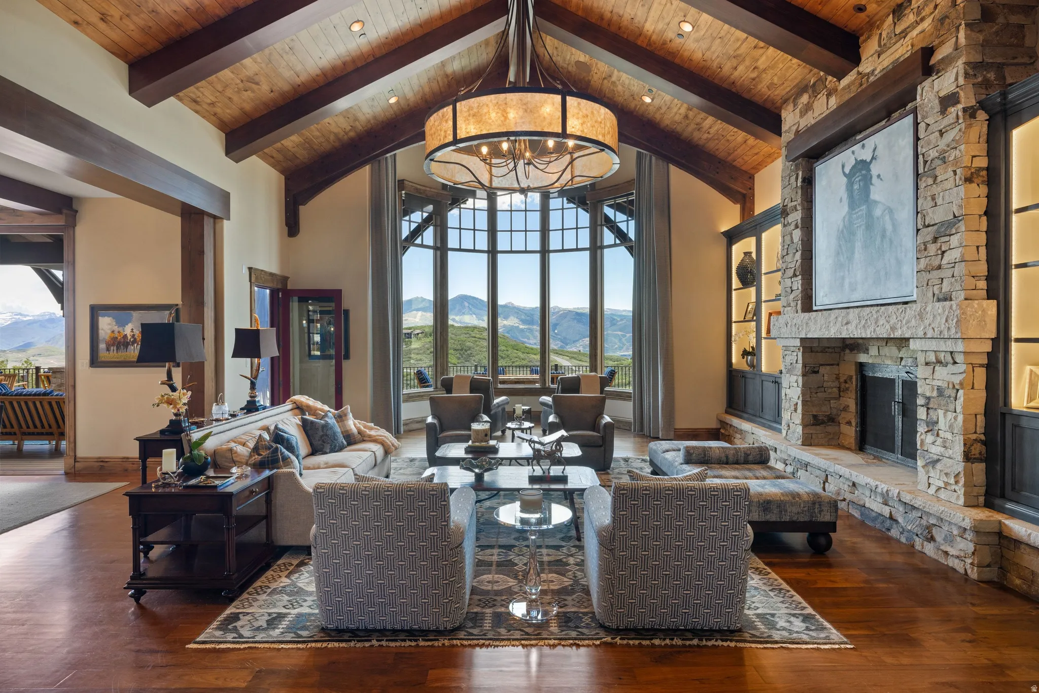 Spacious living area featuring a vaulted wood ceiling with exposed beams, a large stone fireplace with built-in shelving, an oversized drum chandelier, and expansive windows showcasing a mountain backdrop