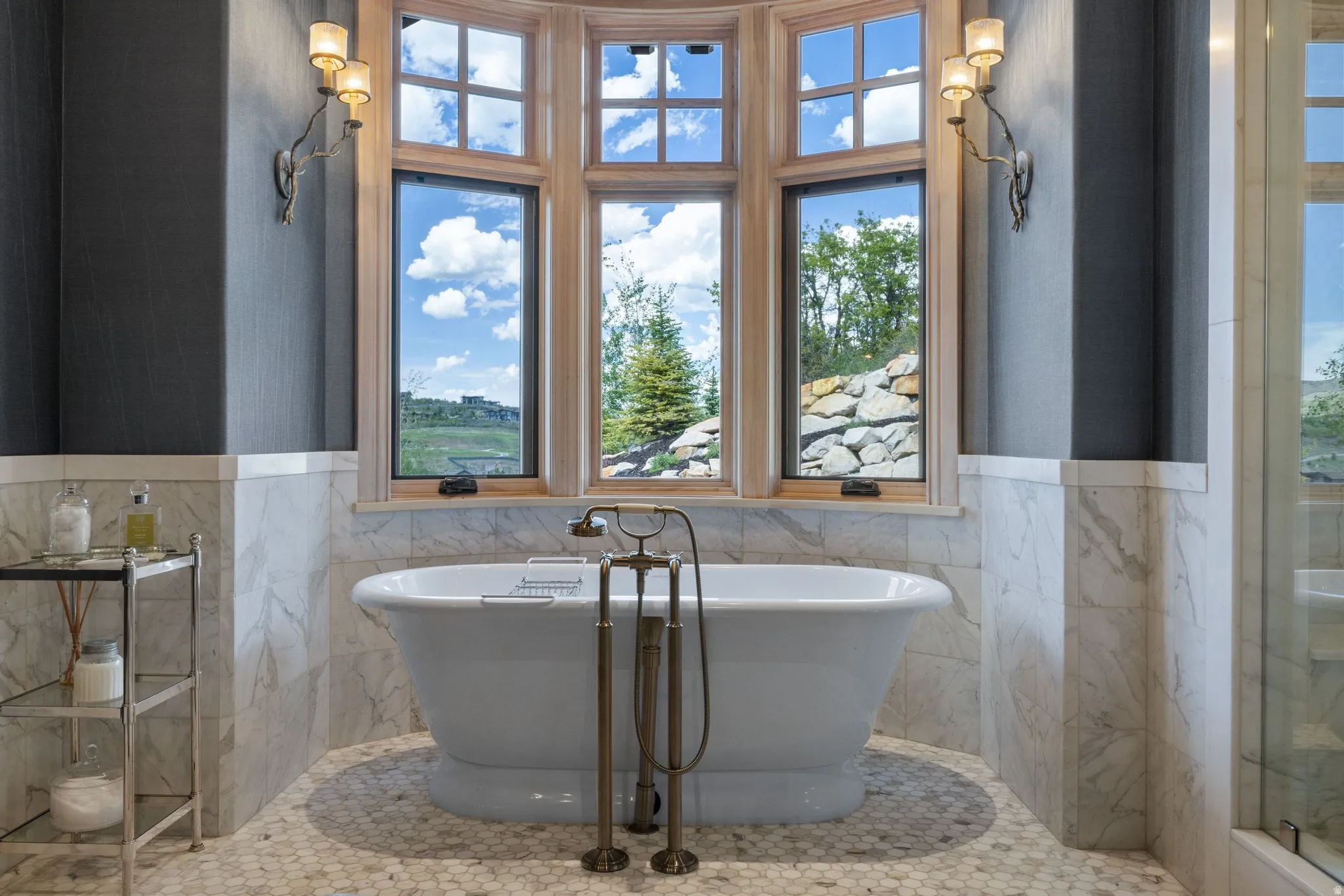 Freestanding tub with brass-finish fixtures, surrounded by marble wainscoting and a multi-pane bay window