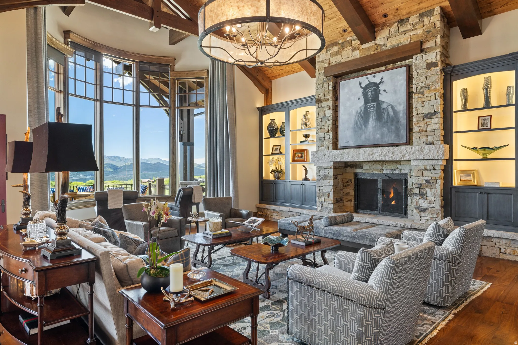 Great room featuring a stone fireplace with hearth seating, custom built-in cabinetry with integrated lighting, exposed wood ceiling beams, and a large arched window with mountain scenery