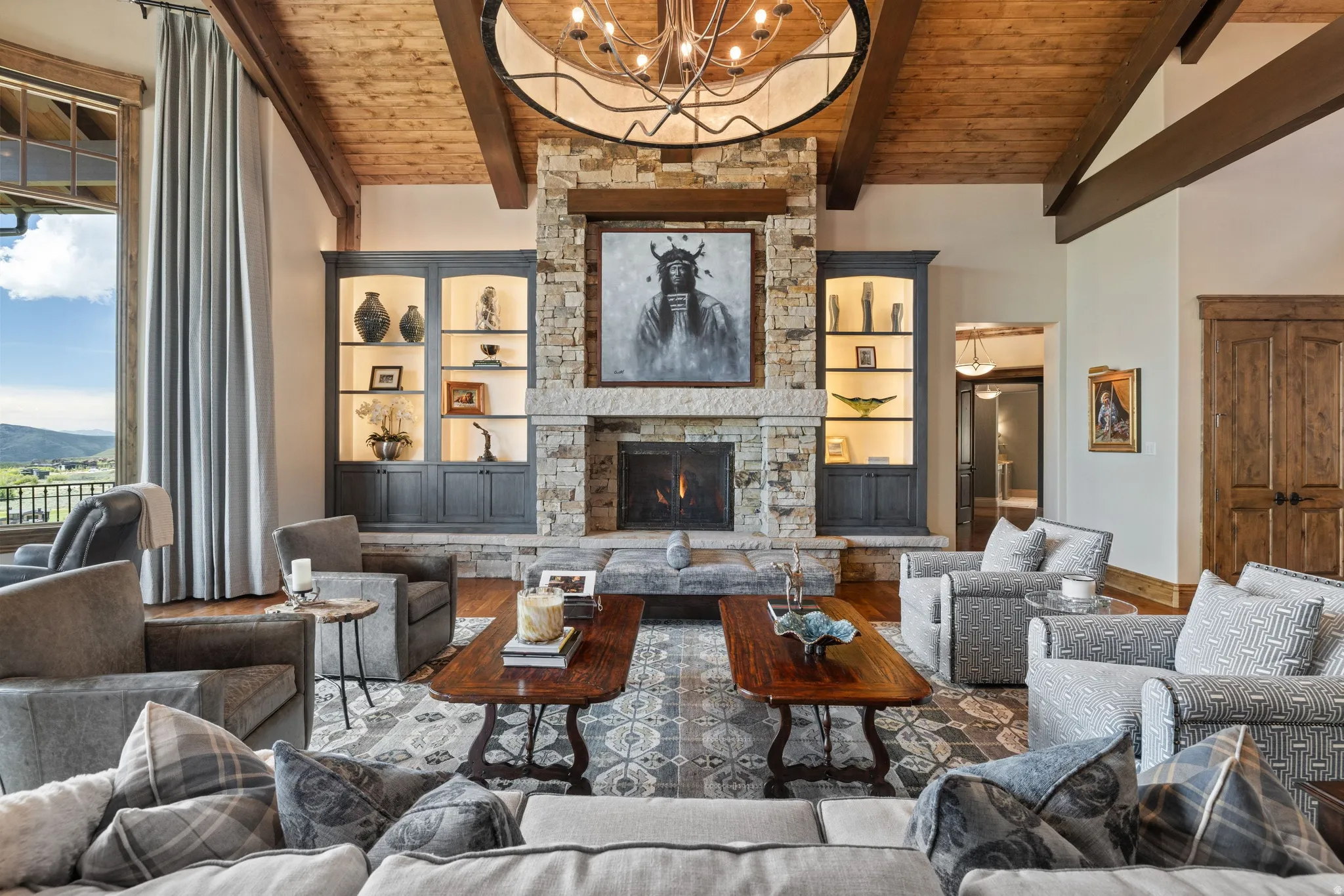 Spacious living area featuring a stone fireplace with a substantial hearth, exposed wood beam ceiling, wood-finish flooring, built-in illuminated display cabinetry, and a large window with a scenic outdoor landscape