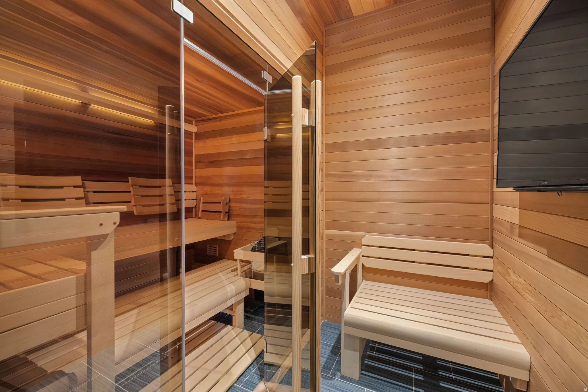 Private sauna featuring clear glass door, multiple slatted wood benches, slatted wood walls and ceiling, and dark tile flooring