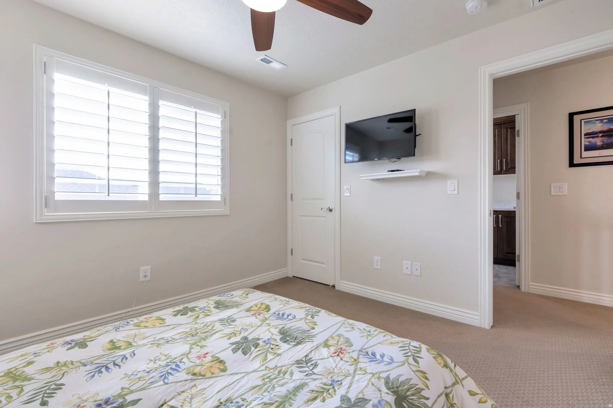 Light-toned wall paint throughout, featuring a ceiling fan with wood-finish blades, a window with white plantation shutters, and light-toned carpet flooring