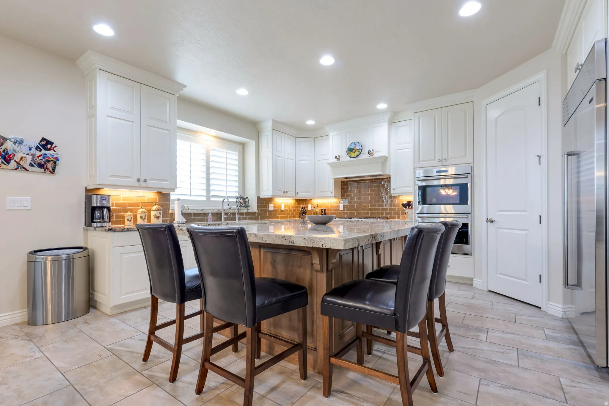 Kitchen featuring a large central island with a light-colored countertop, white shaker-style cabinetry, stainless steel appliances including a built-in oven and refrigerator, a textured tile backsplash, and light-toned tile flooring