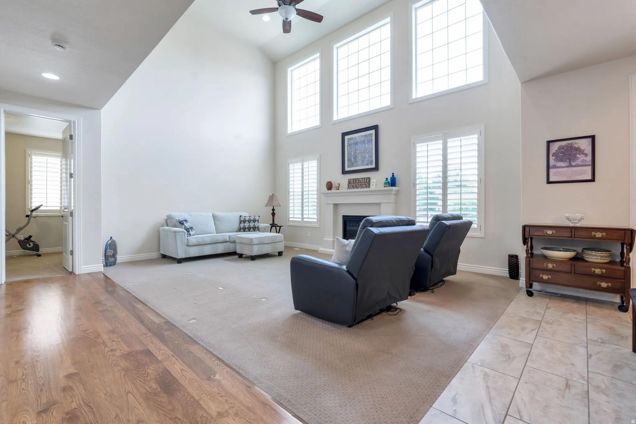 Spacious living area featuring a vaulted ceiling, multiple large windows with plantation shutters, a ceiling fan, and a fireplace with a mantel
