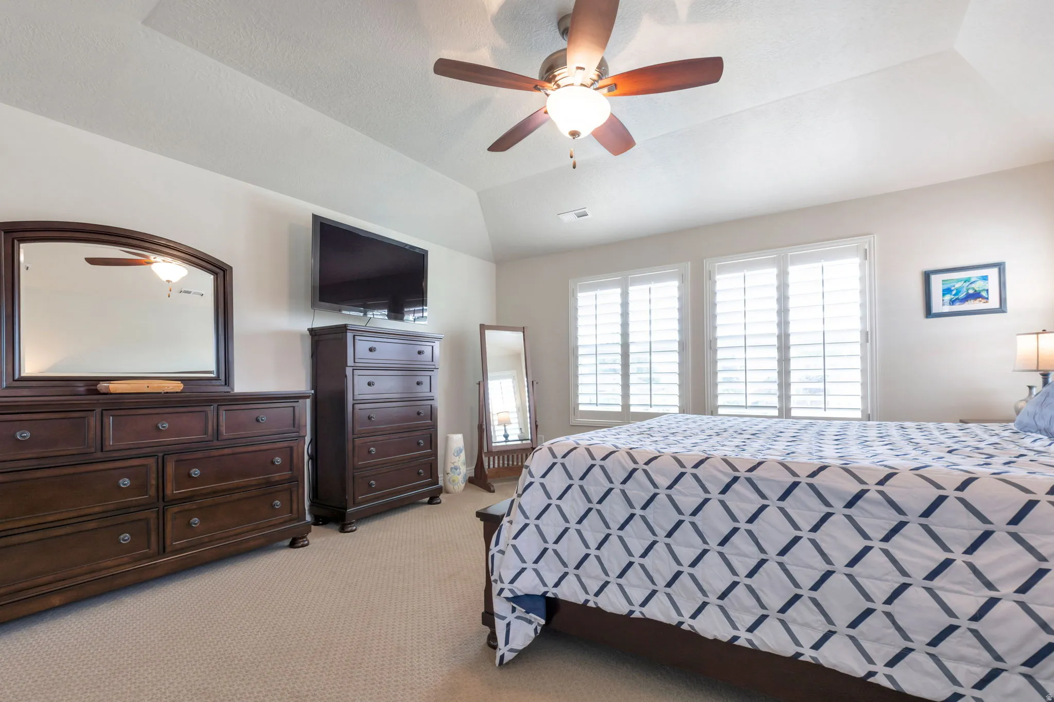 Spacious room featuring vaulted ceilings, light-colored carpeting, two large windows with plantation shutters, and a ceiling fan with integrated lighting