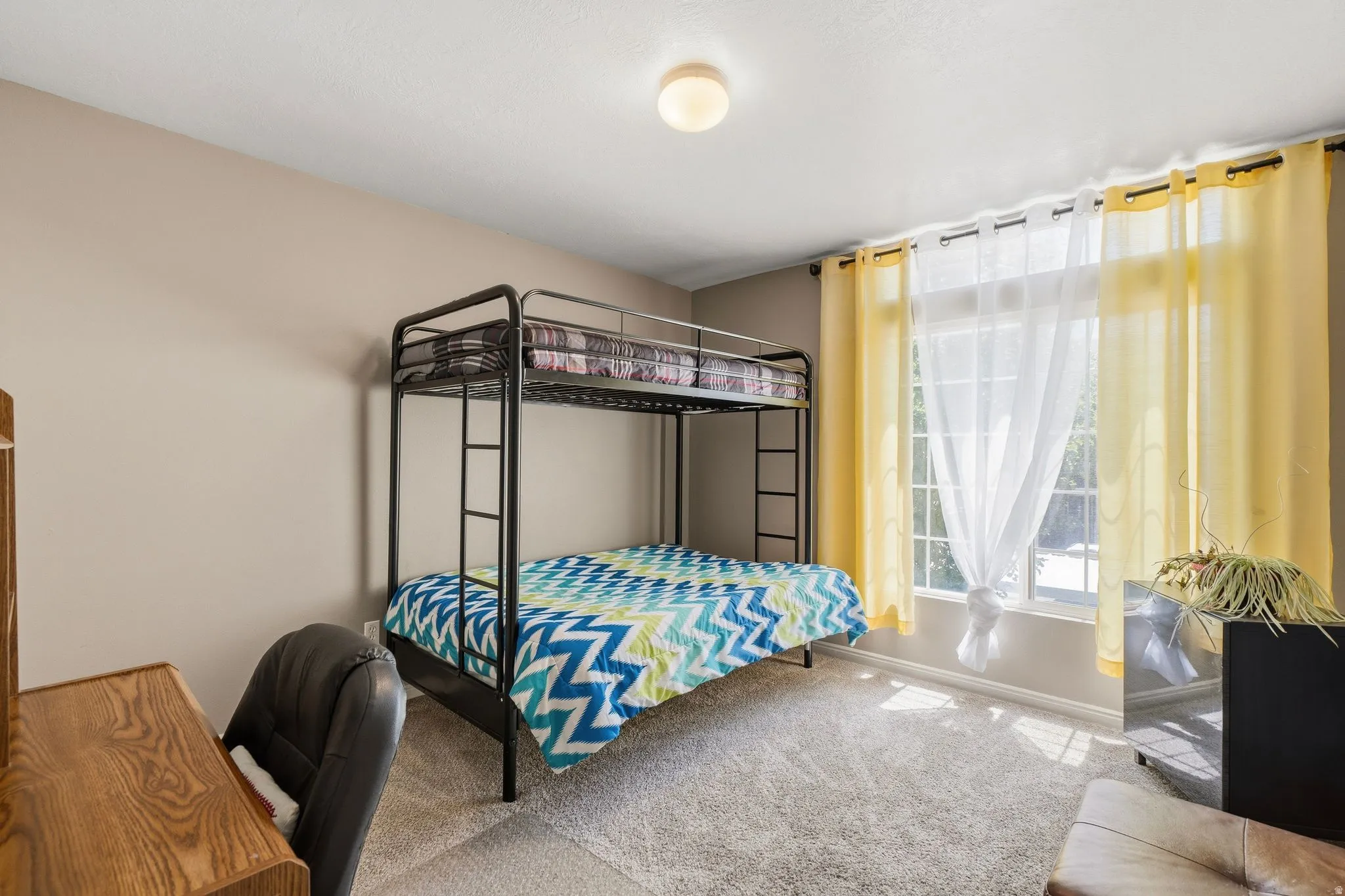 Bedroom with plush carpet flooring, neutral painted walls, and a large window featuring sheer and yellow drapes