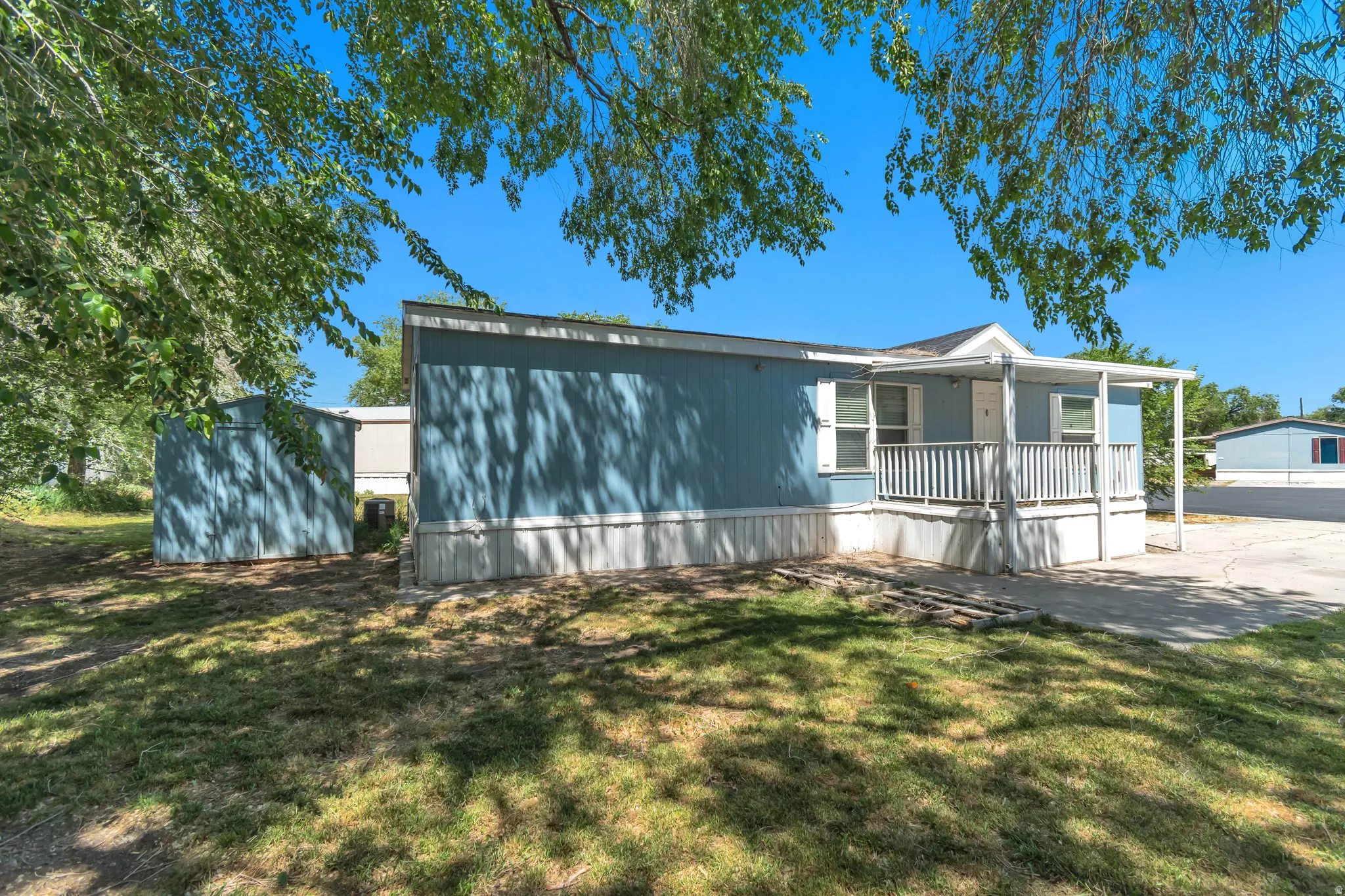 Blue manufactured home featuring a covered front porch with white railing, light-colored trim, and a separate blue utility shed