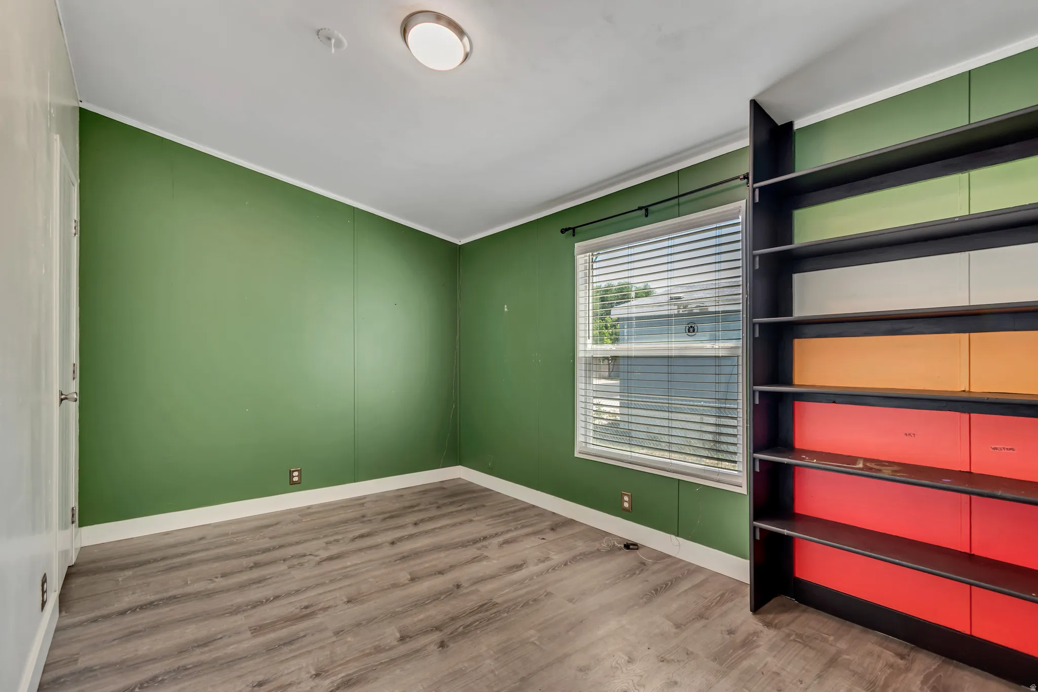 Room featuring wood-finish flooring, a window with blinds, and a built-in shelving unit with colored backing