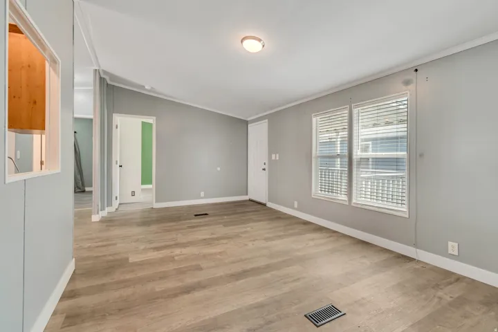 Spacious room featuring wood-finish flooring, two windows with blinds, a white entry door, and a pass-through window with wood-paneled interior