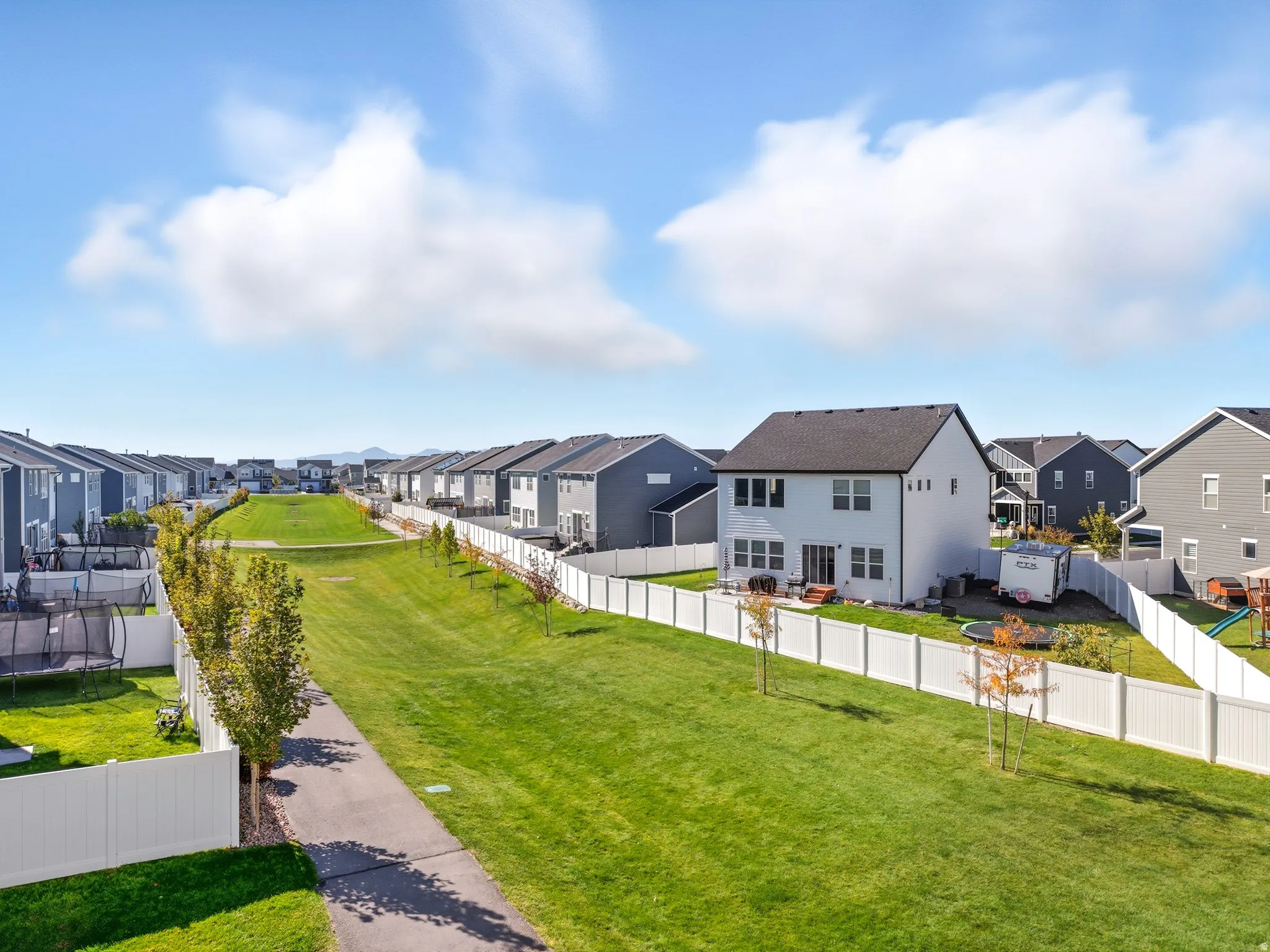 Expansive green space featuring a paved walking path, mature trees, and a white vinyl fence