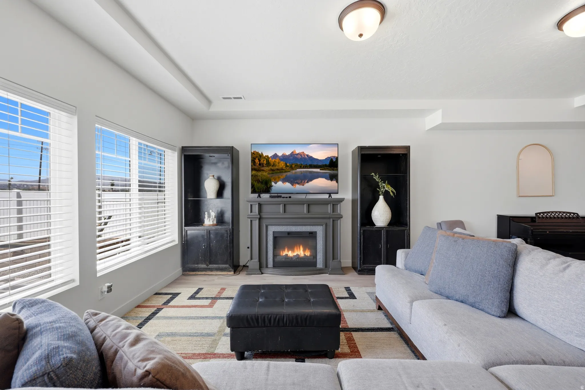 Spacious living area featuring a built-in fireplace with mantel, recessed ceiling details, multiple windows with blinds, and wood-finish flooring