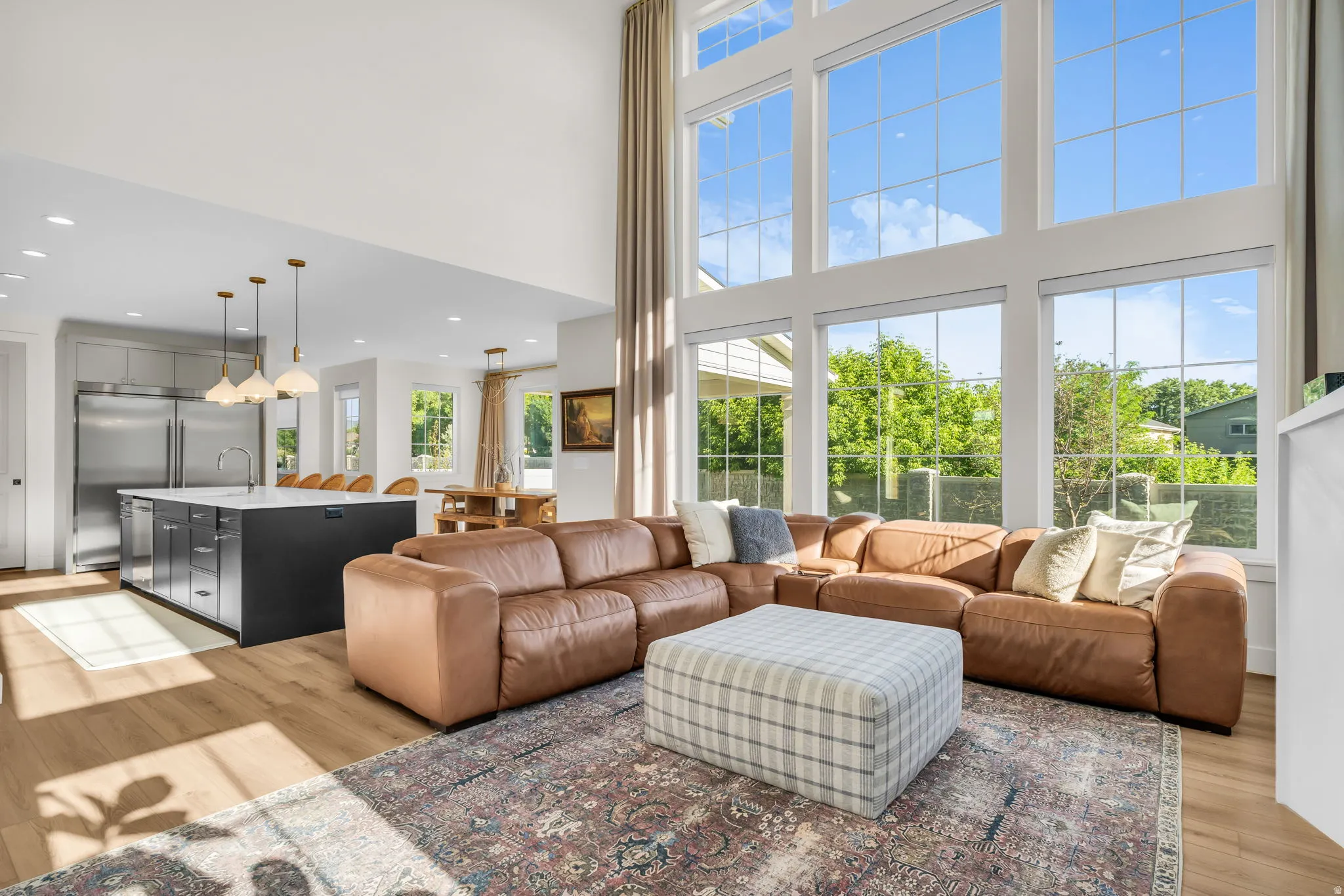 Two-story living area with expansive window wall, wood-finish flooring, and a large open-concept kitchen featuring a dark island with seating and stainless steel appliances
