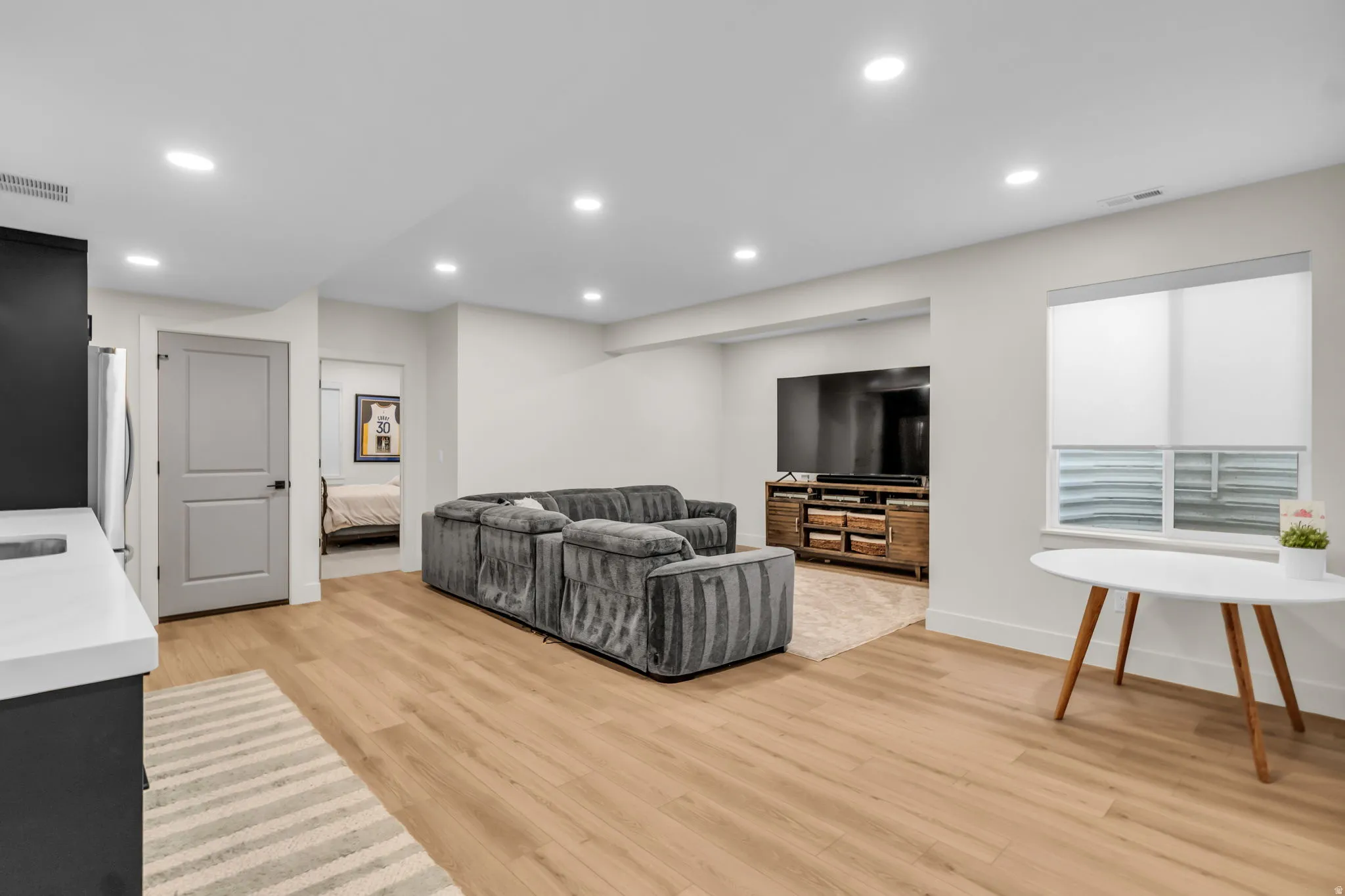 Open-concept living space featuring light wood-finish flooring, recessed lighting, a built-in wet bar with white countertop, and a window with roller shades