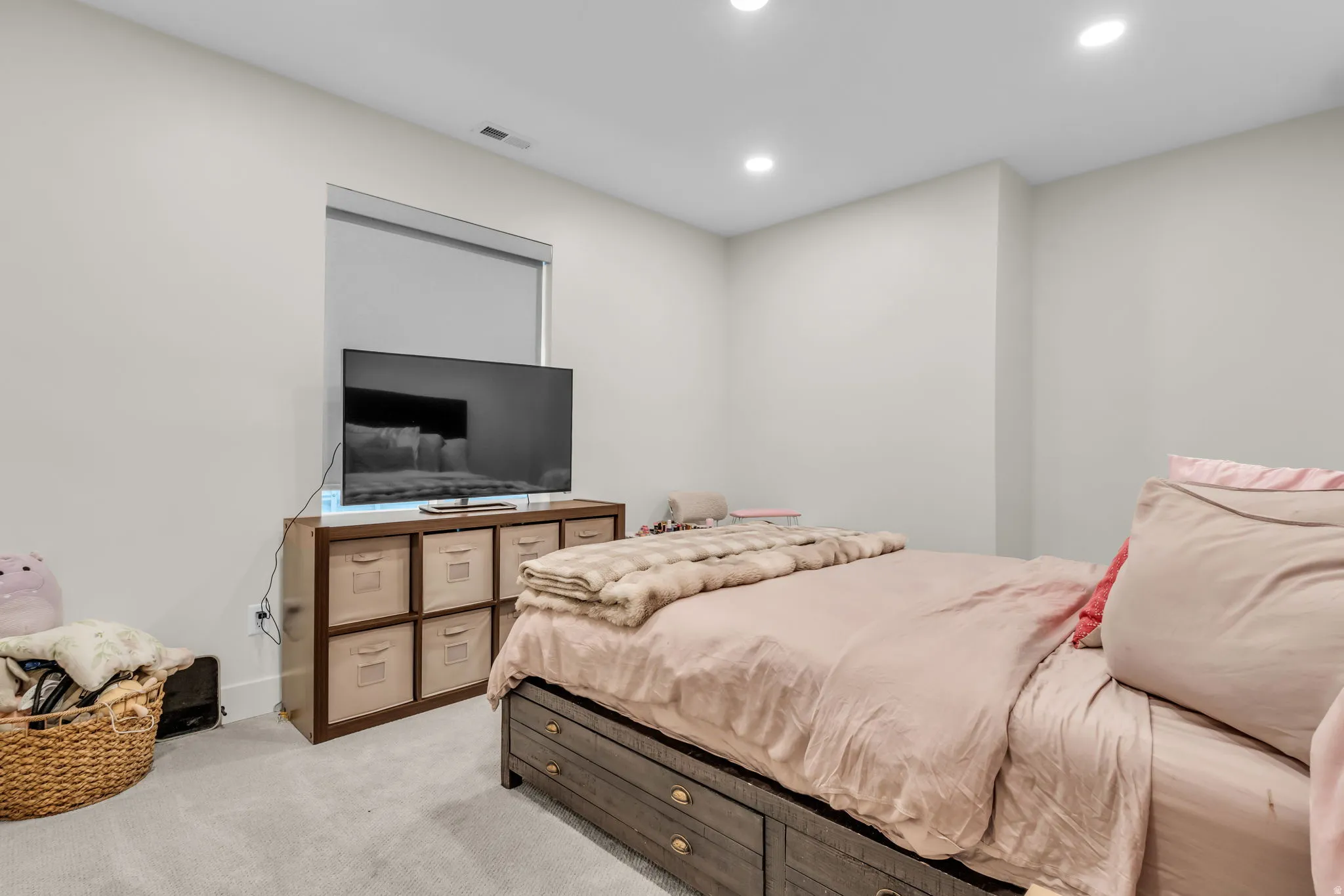 Recessed lighting illuminates the space, featuring light gray walls, a window with a roller shade, and plush gray carpeting