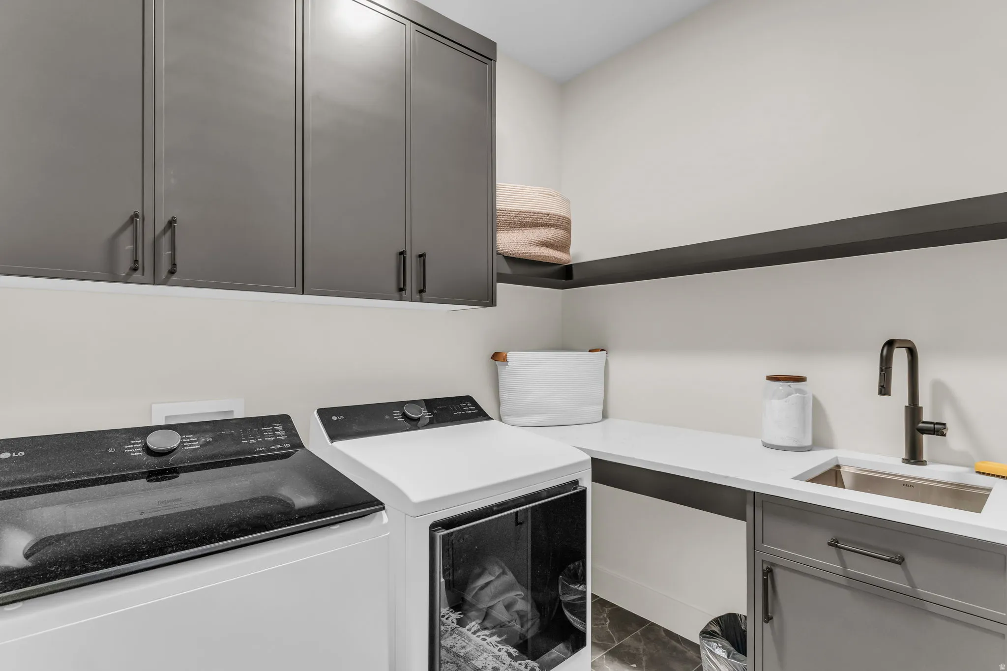 Utility room featuring dark gray cabinetry, white countertops, a stainless steel sink with a dark-finish faucet, an open dark-finish shelf, and dark tile flooring