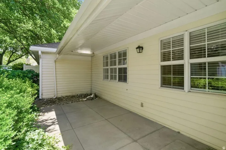 Covered concrete patio with light yellow siding, multiple windows with blinds, an exterior wall sconce, and surrounding landscaping