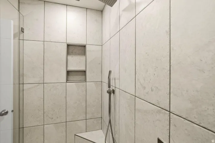 Tiled shower enclosure featuring light-toned large format tiles, a built-in recessed niche, a corner bench, a rainfall shower head, and a handheld shower fixture