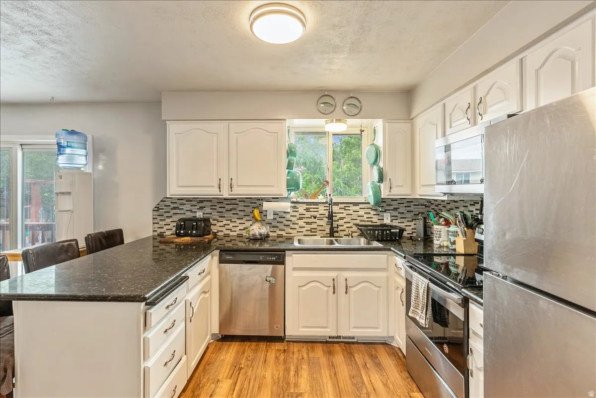 Kitchen featuring white cabinetry with dark hardware, dark countertops, a mosaic tile backsplash, stainless steel appliances, and wood-finish flooring