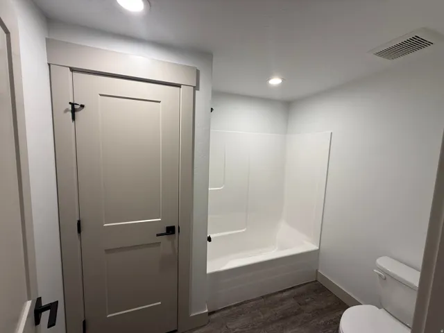 Bathroom featuring a white bathtub/shower combination, a toilet, wood-finish flooring, and recessed ceiling lighting